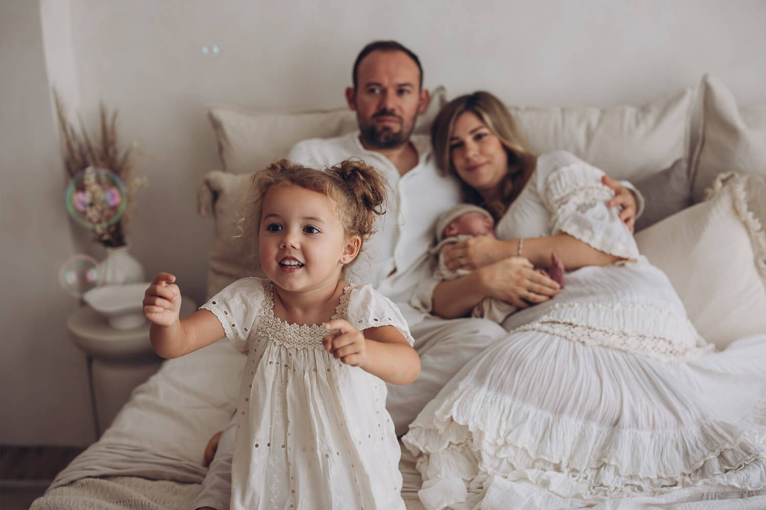 Authentische Familienreportage in Peine – Kleinkind spielt im Vordergrund während Eltern ihr Neugeborenes im Bett halten