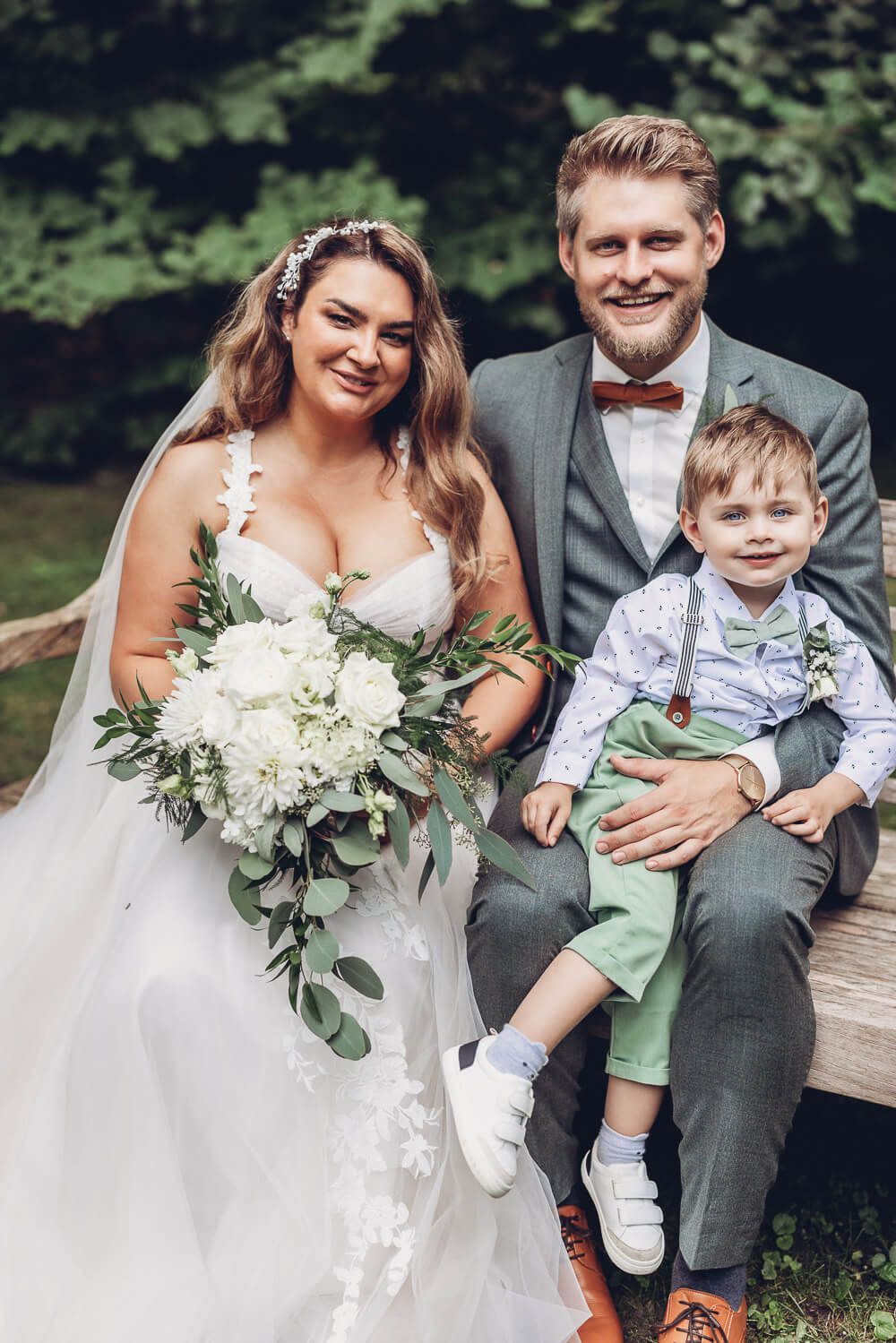 Natürliches Familienportrait von Brautpaar und Kind auf einer Holzbank im Grünen, fotografiert von einem Fotografen in Hannover