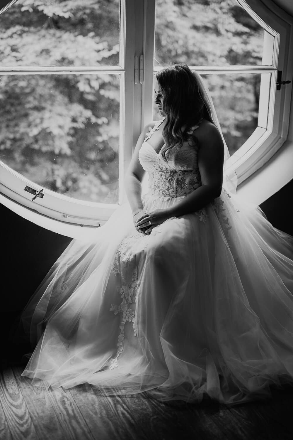 Künstlerisches Schwarz-Weiß-Portrait einer sitzenden Braut am Fenster in ihrem Hochzeitskleid, fotografiert von einer Fotografin in Celle