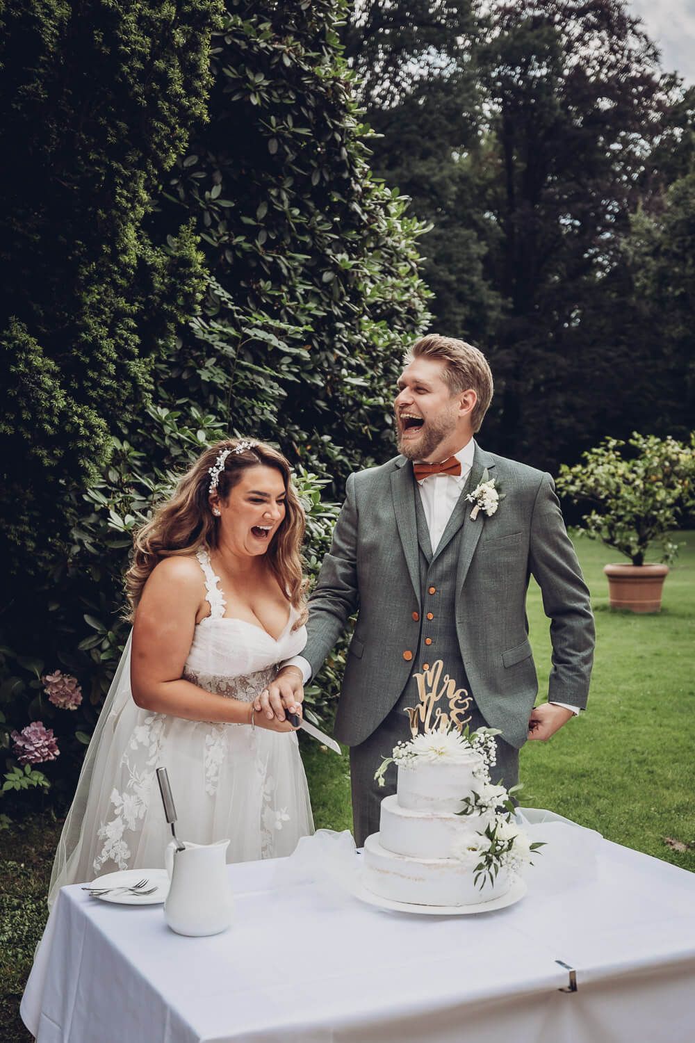 Fröhliches Brautpaar schneidet gemeinsam die Hochzeitstorte im Garten an, aufgenommen von einer Hochzeitsfotografin in Hameln