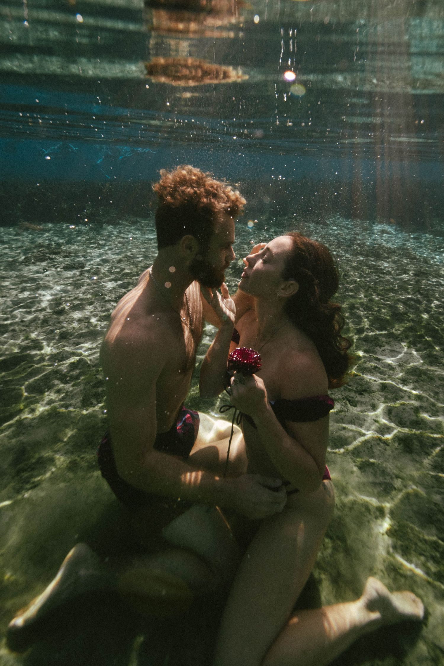 Artistic underwater photograph showing two people embracing in clear turquoise water against sunlit reflections.