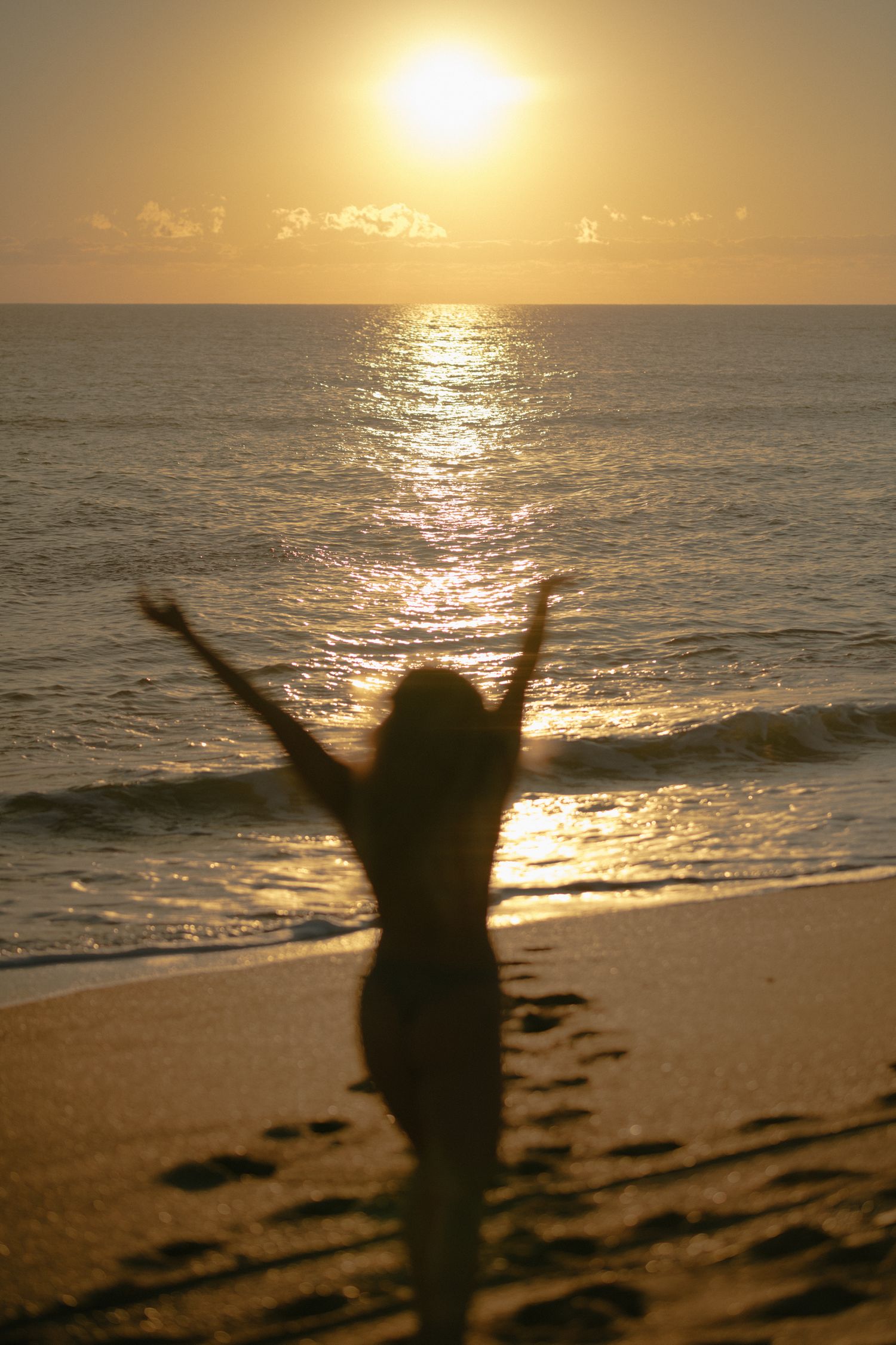 Silhouette of a person with raised arms celebrating on a beach at sunset with golden reflections on the ocean.