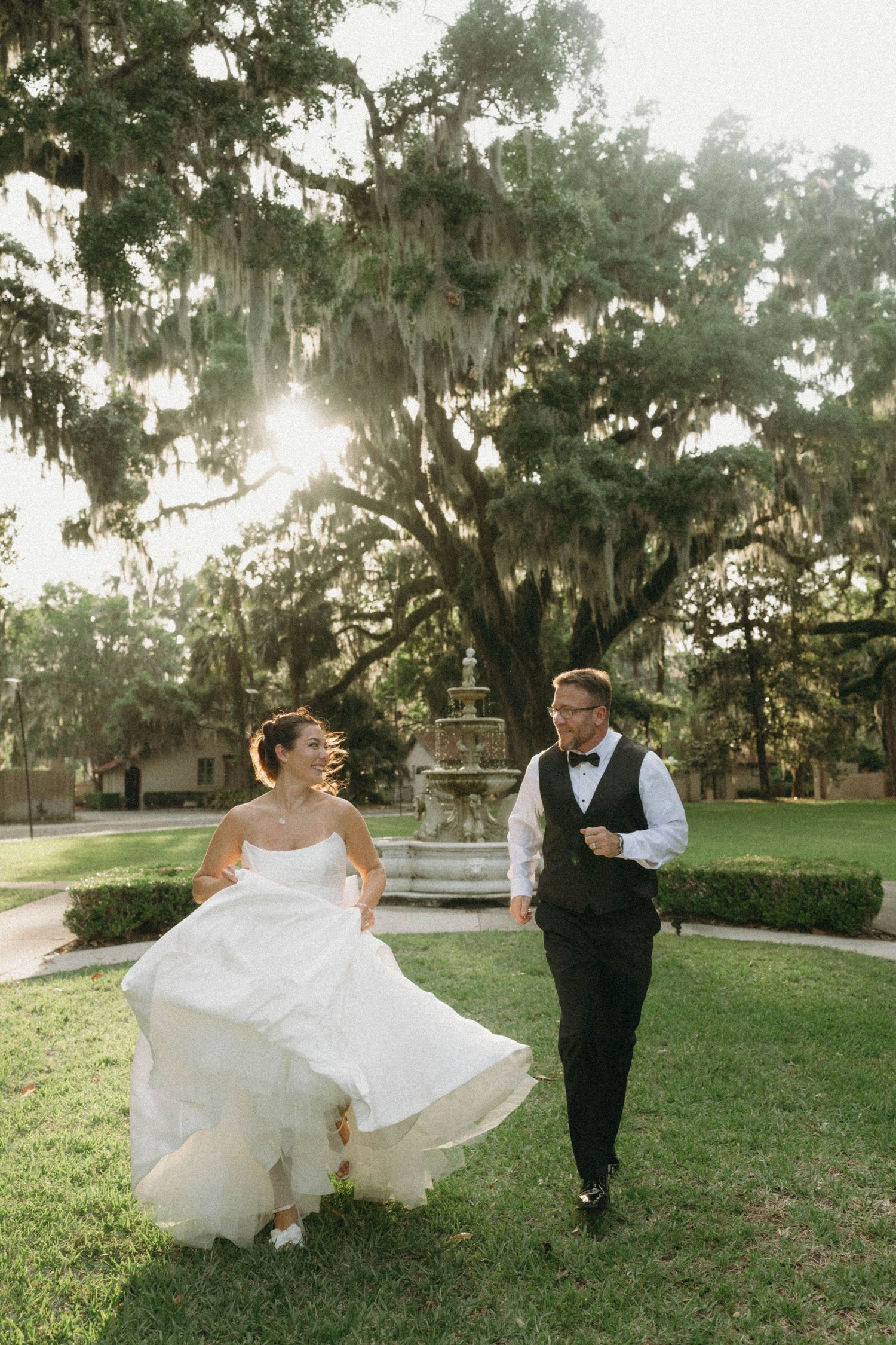 A couple runs playfully across a sunlit lawn with Spanish moss draped trees creating a romantic Southern backdrop.