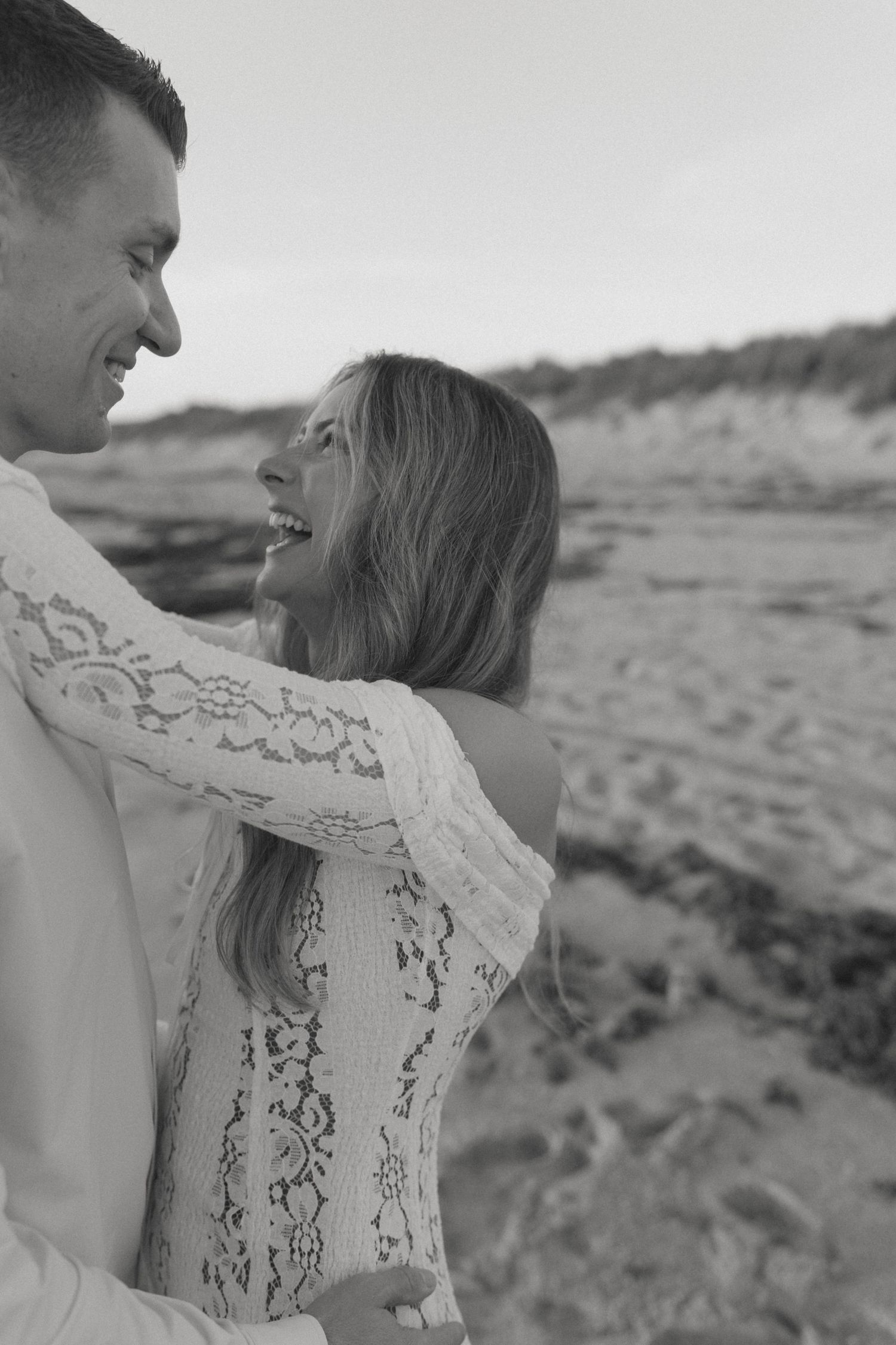 Black and white photo of a smiling couple embracing on a sandy beach, woman wearing lace dress.