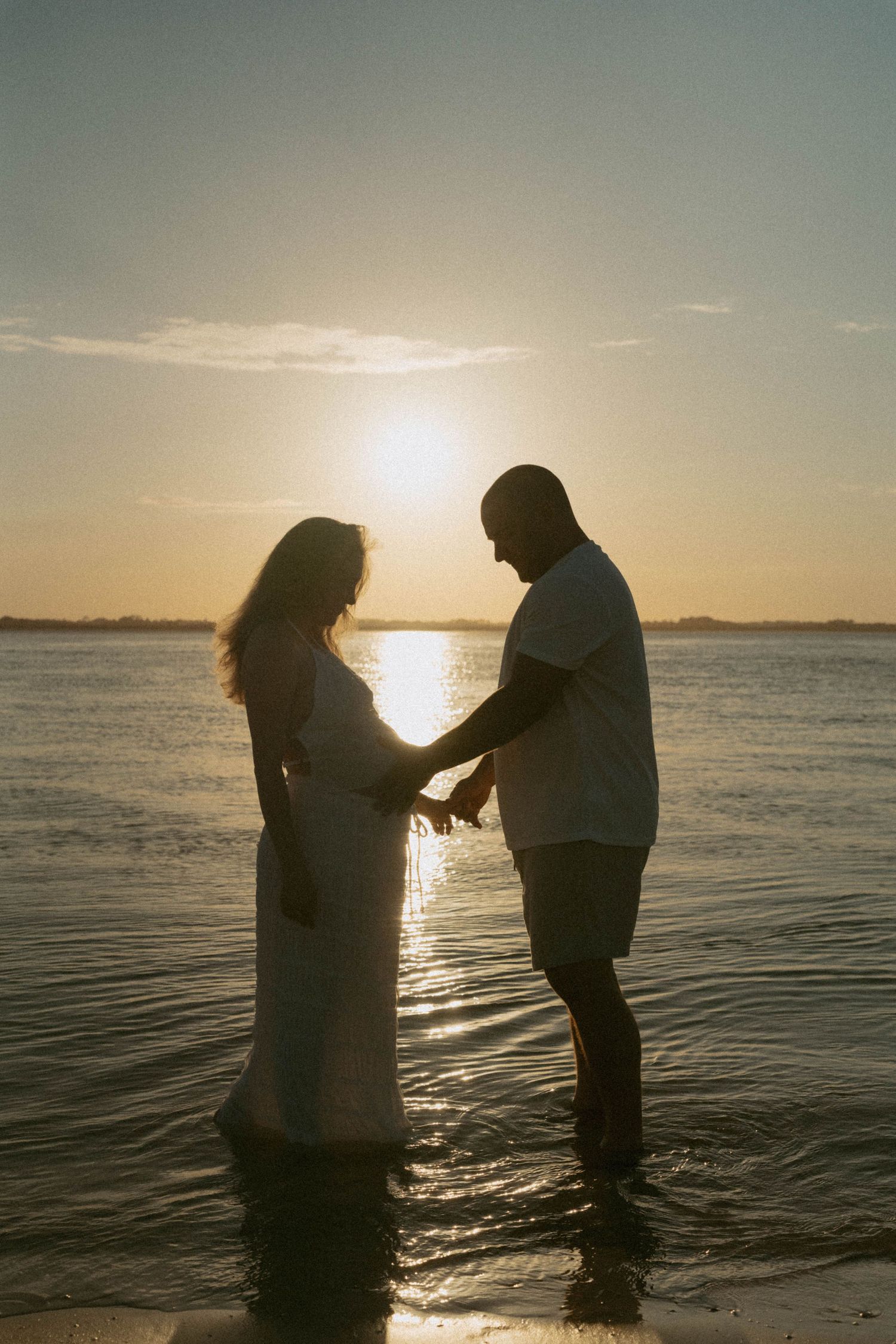 Silhouette of a pregnant couple holding hands in shallow water during a golden sunset at the beach.