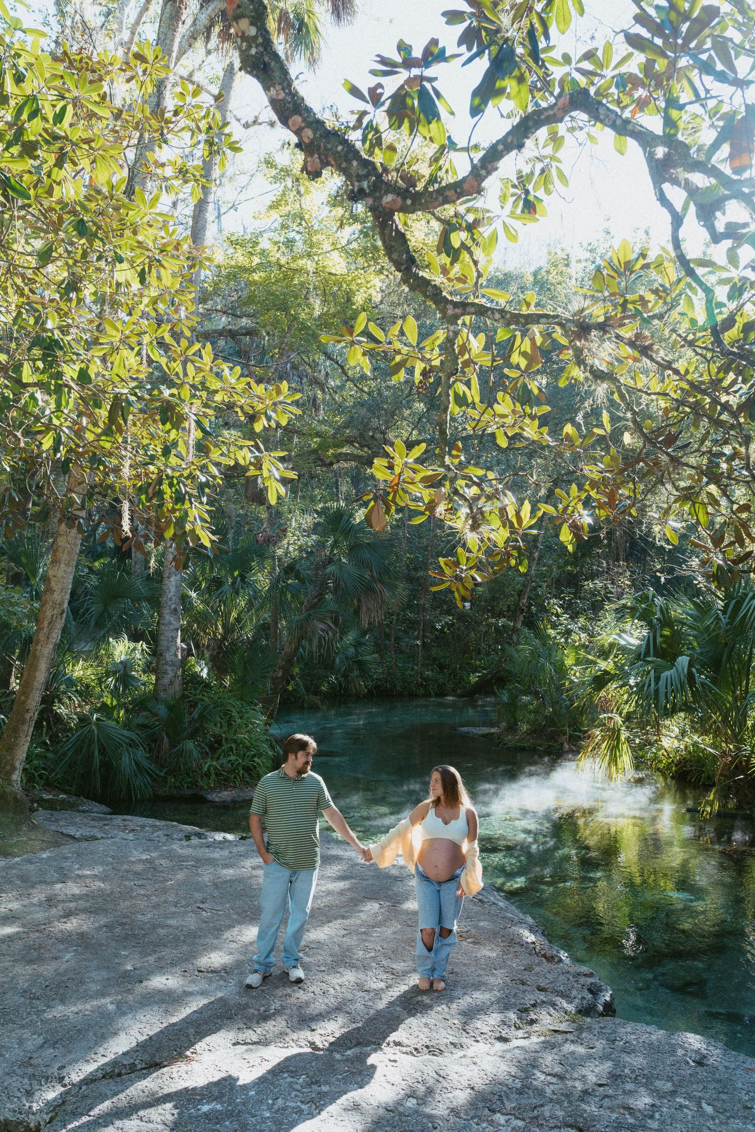 People walking along a scenic nature trail surrounded by lush tropical foliage near a waterway on a sunny day.