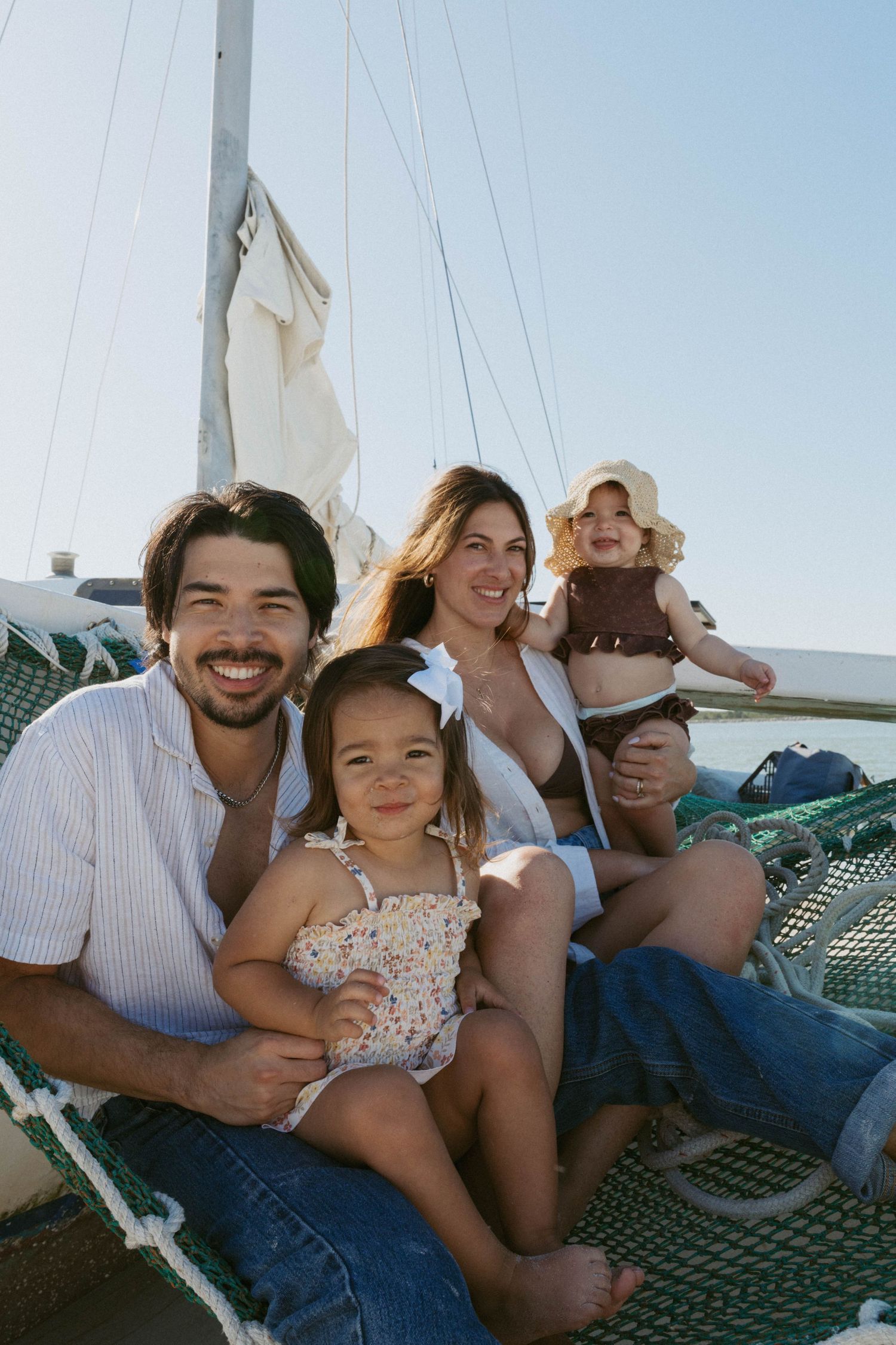 Happy family with two young children sitting on a sailboat deck on a sunny day, smiling outdoors.