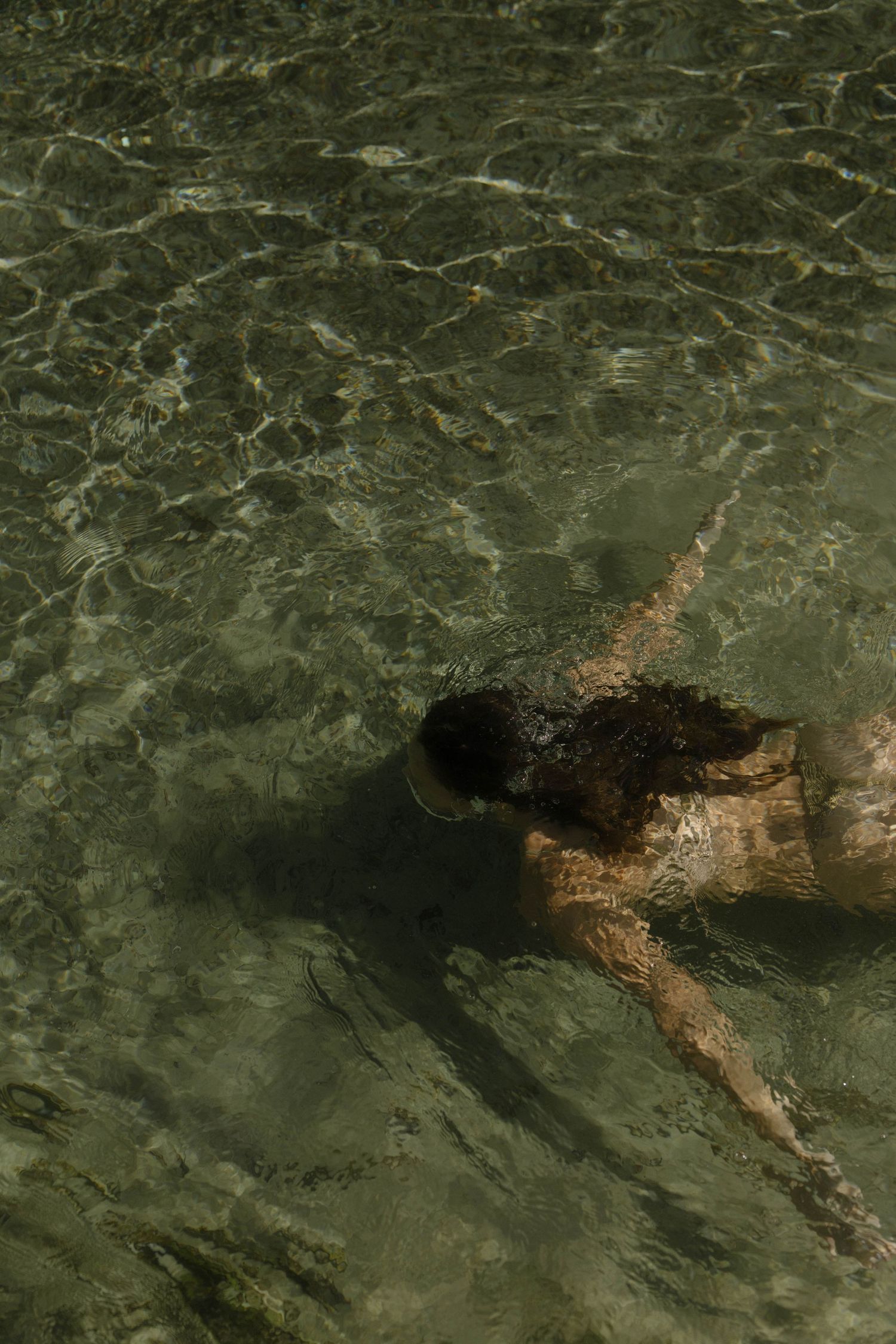 Person swimming underwater in clear shallow green ocean water with visible sandy bottom.