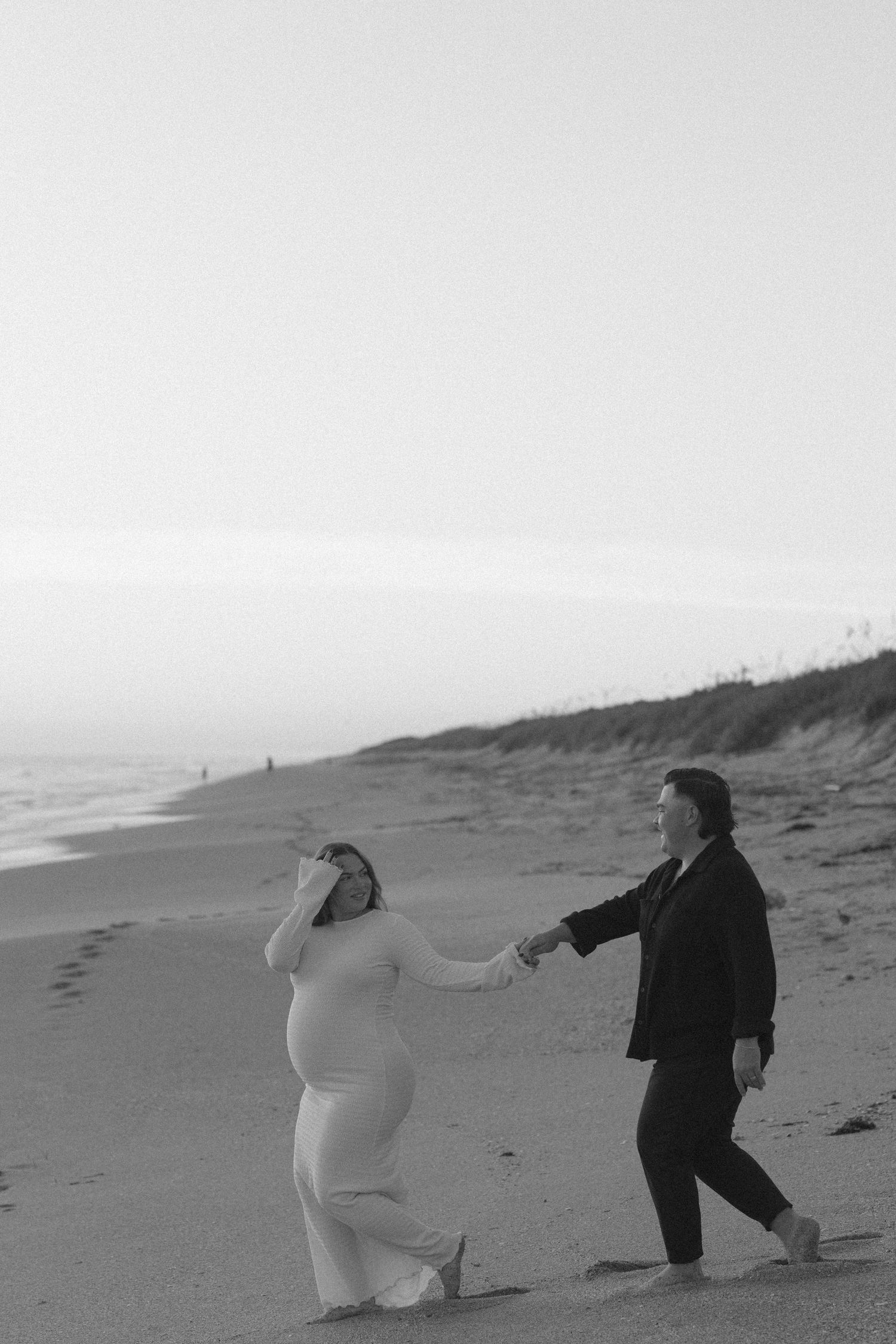 Two people walk together holding hands along a misty beach shoreline in black and white.