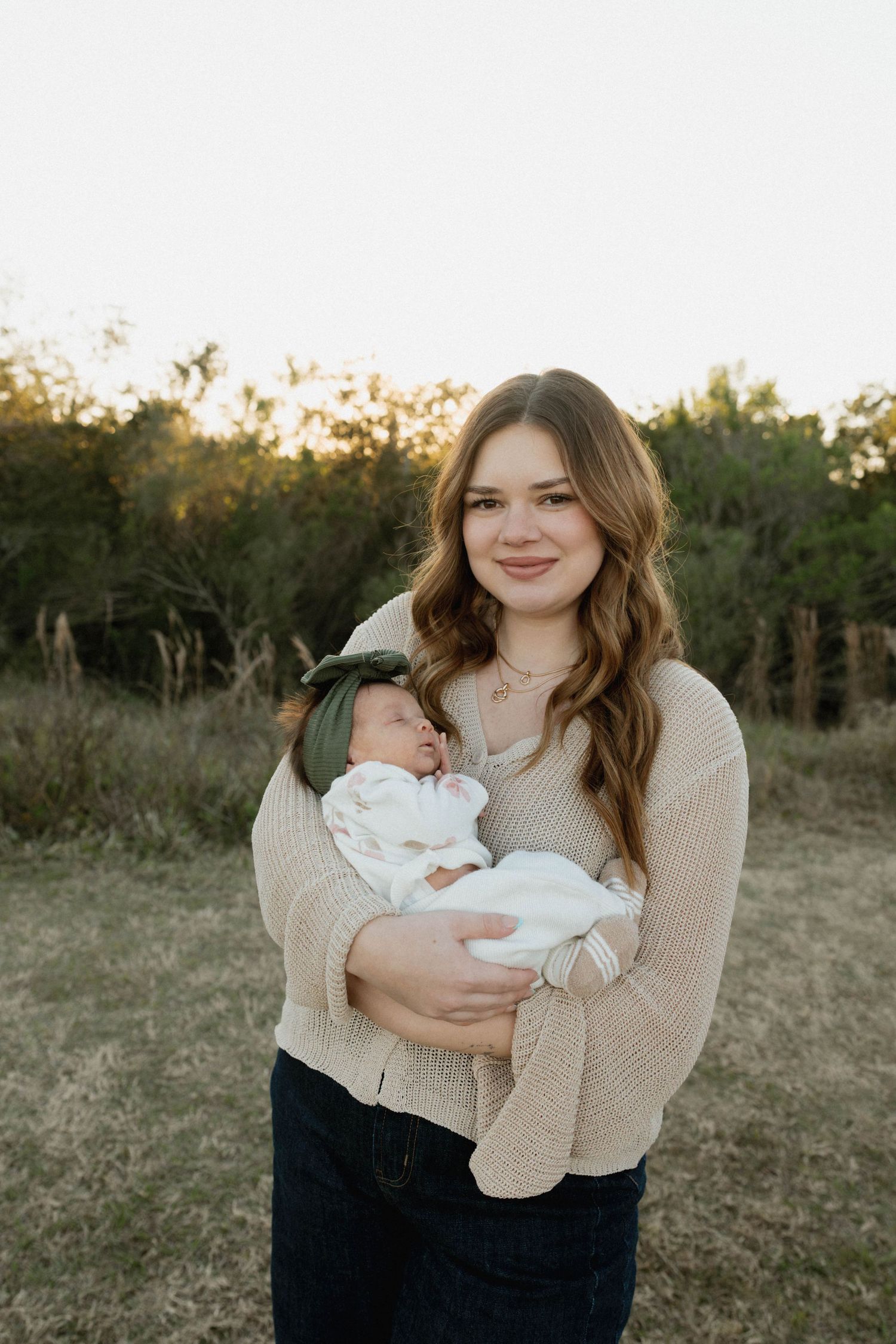 A parent in a beige sweater holds a newborn baby in white clothing outdoors during golden hour.
