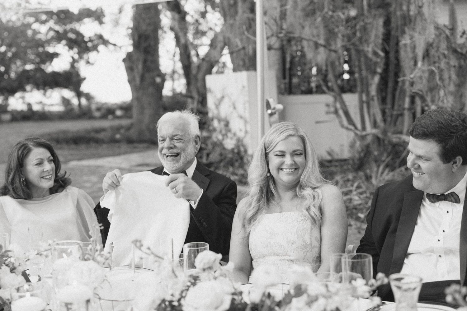 Black and white photo of a bride and groom laughing with family during wedding reception dinner toast.
