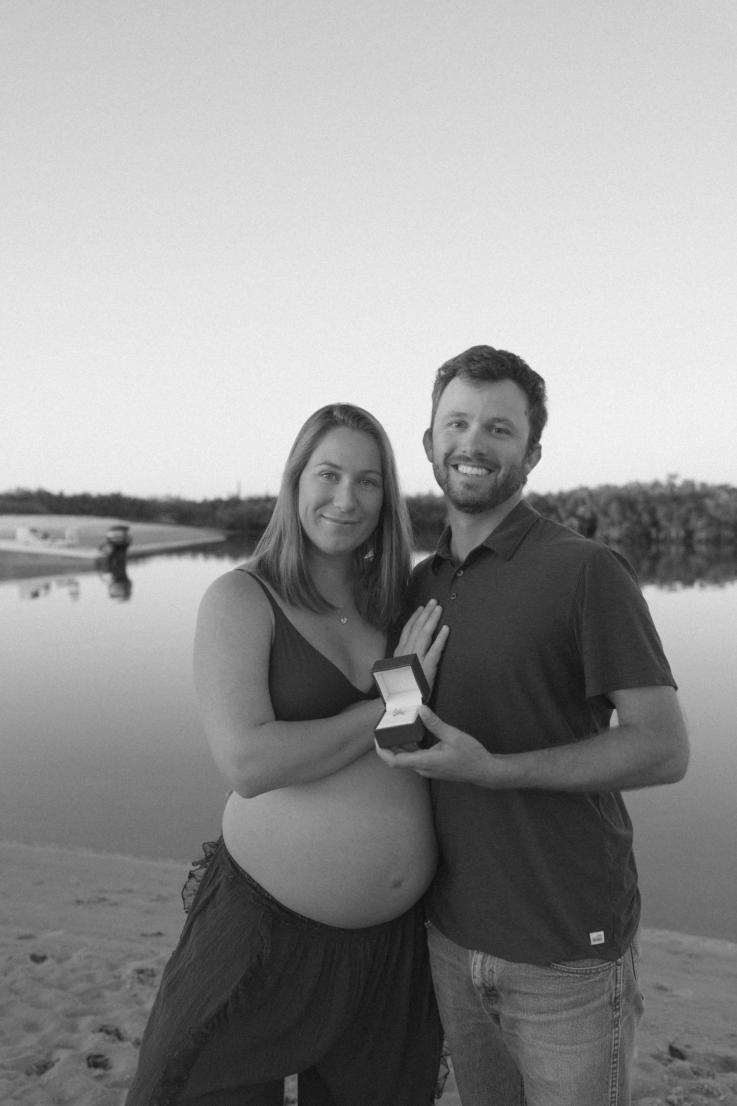 Black and white photo of a smiling pregnant couple holding a ring box by a serene river beach.