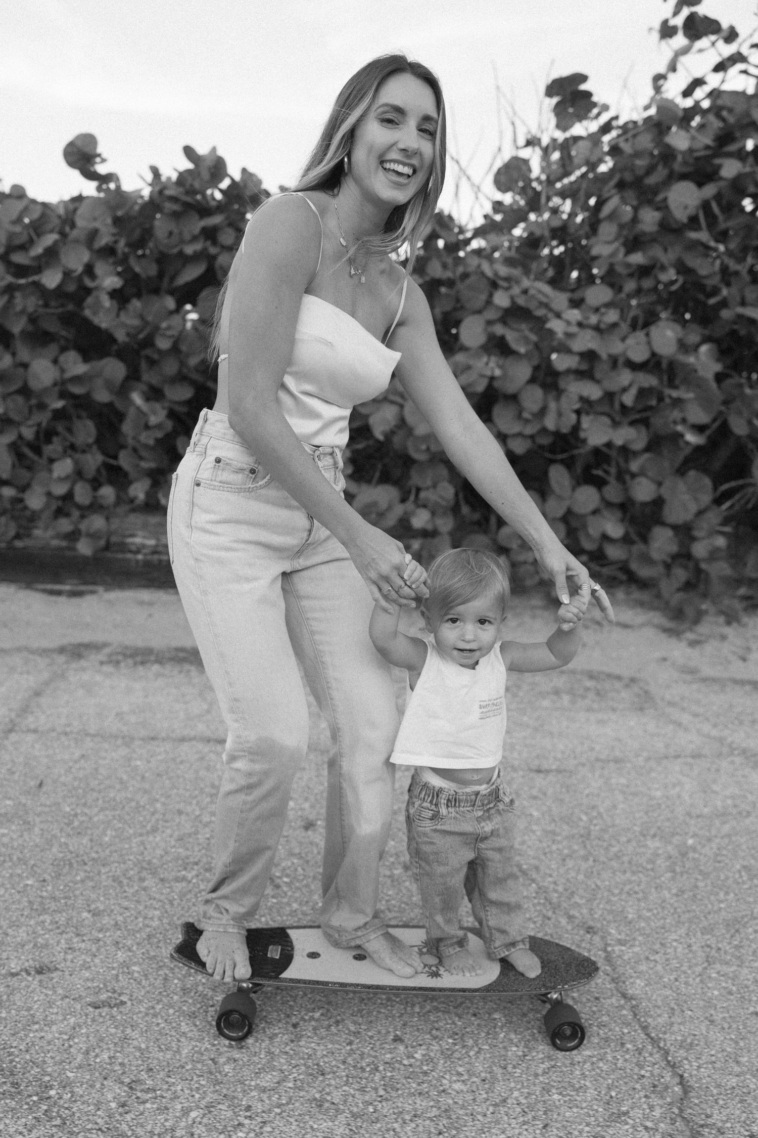 Black and white photo sequence showing a parent teaching a small child how to skateboard on a paved surface.