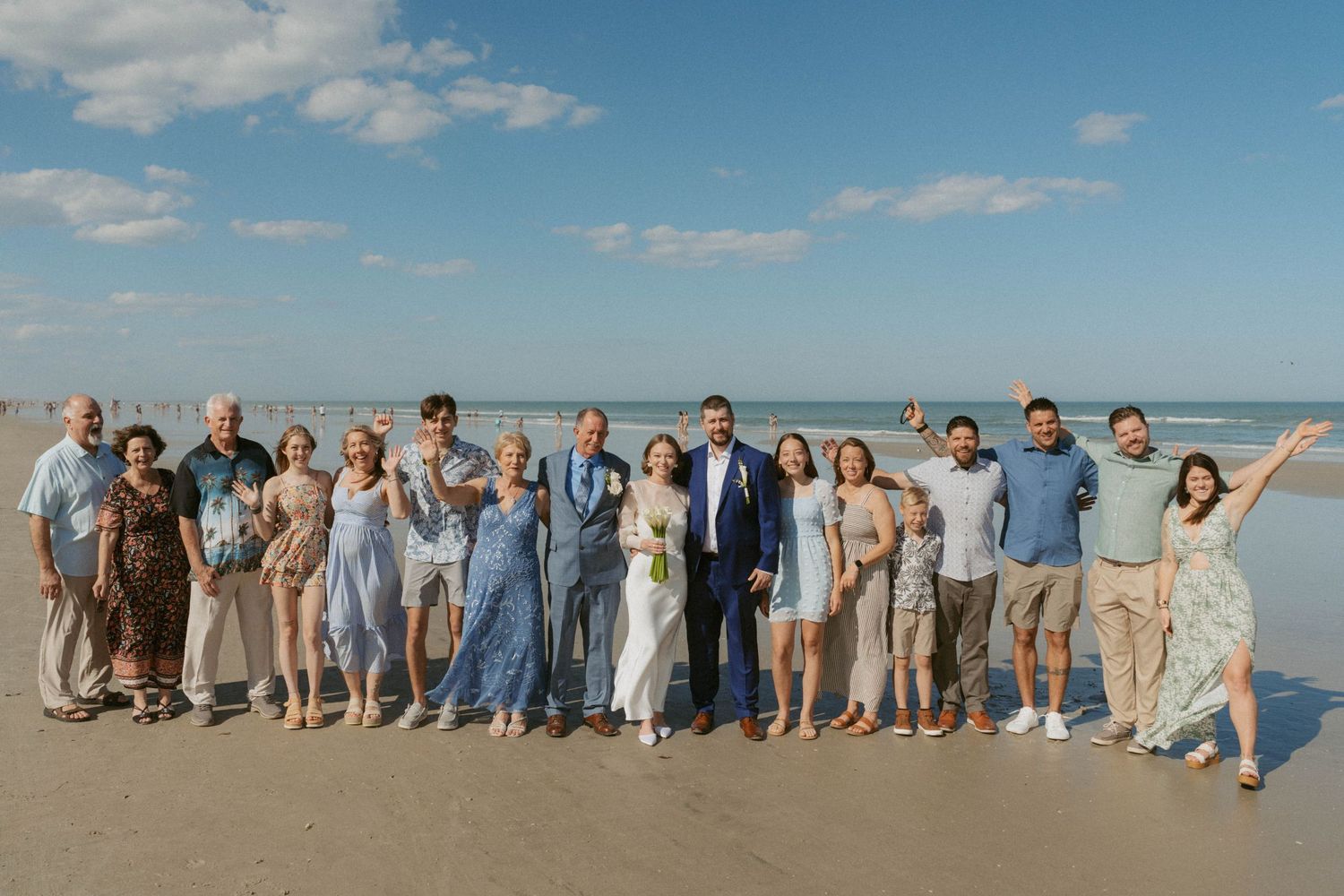 Large group of people gathered on a sunny beach wearing casual attire and posing together for a photo.