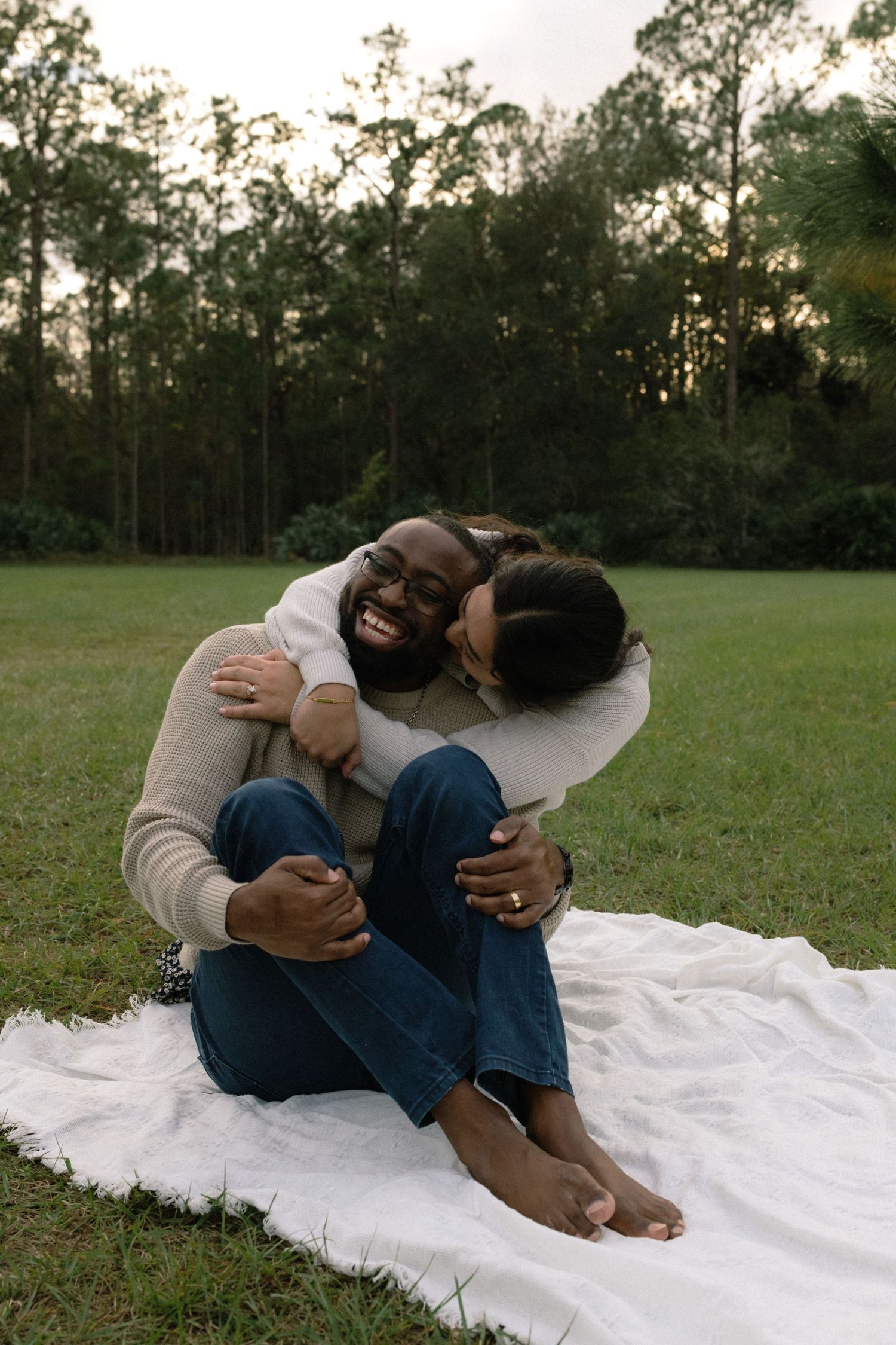 A couple shares a tender moment while sitting on a white blanket in a grassy field with tall trees at dusk.