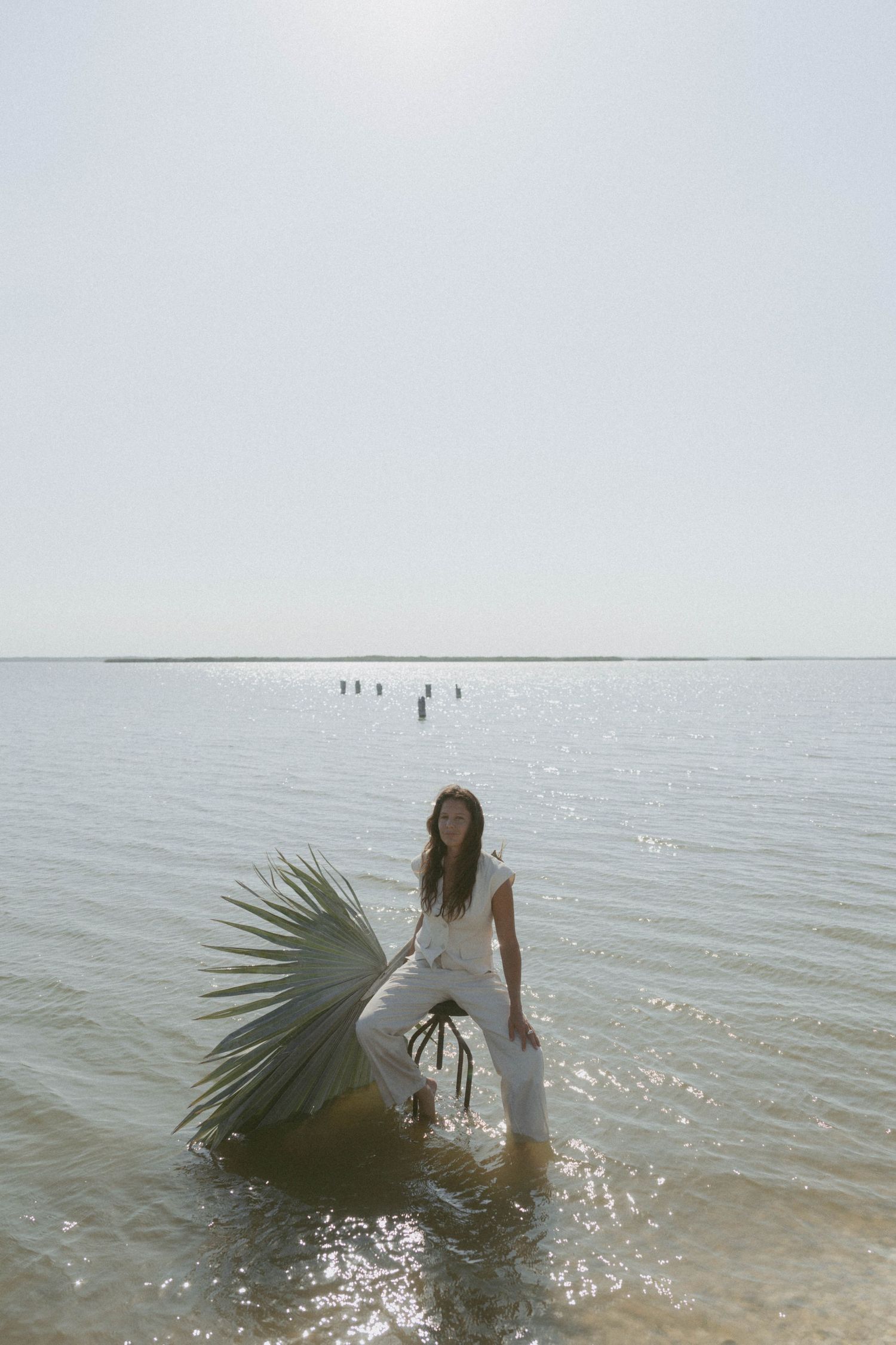 Woman in white outfit holding a palm leaf standing in shallow ocean water on a bright sunny day.