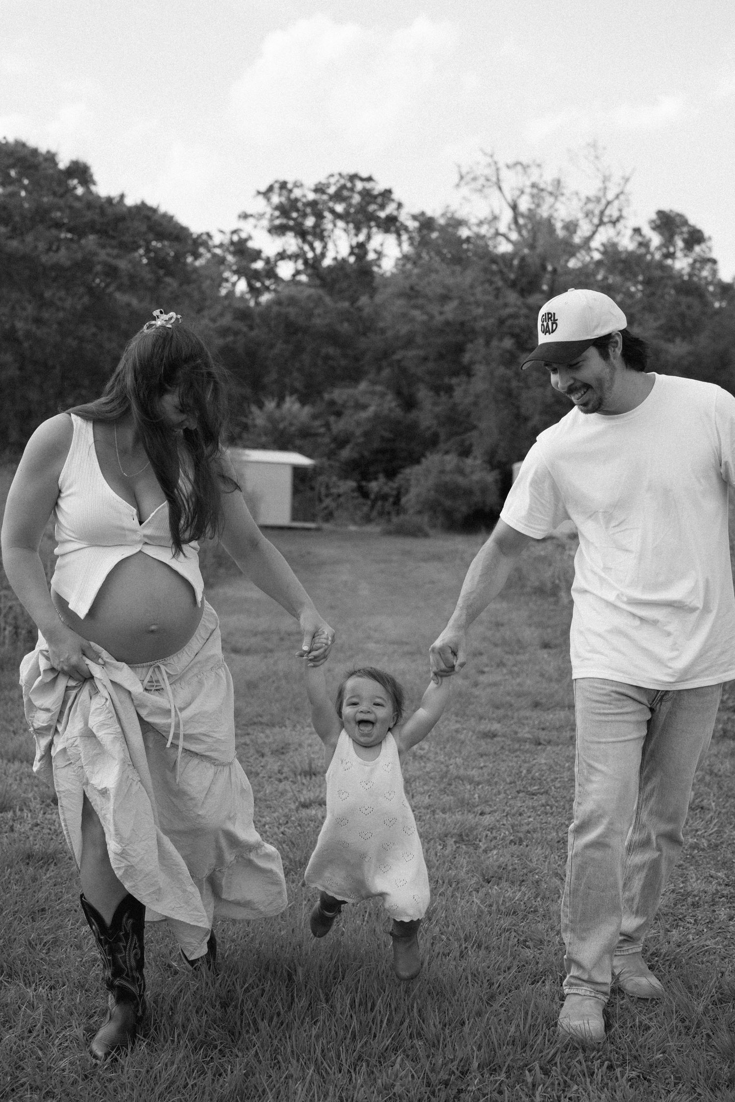 Black and white photo of a joyful moment captured outdoors showing an expectant parent and partner holding hands with their toddler.