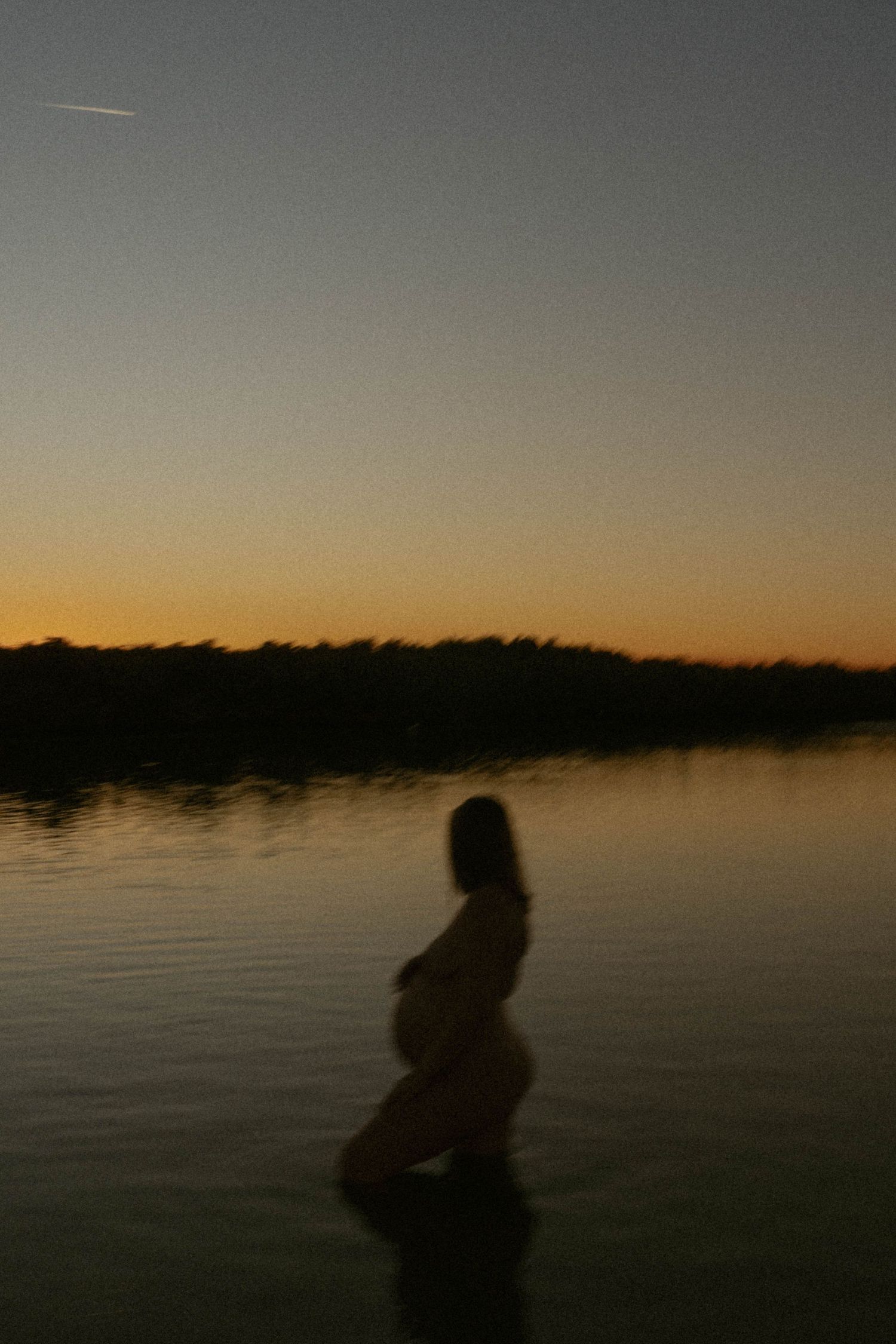 Silhouettes of figures stand against a tranquil lake at sunset with a warm orange glow on the horizon.