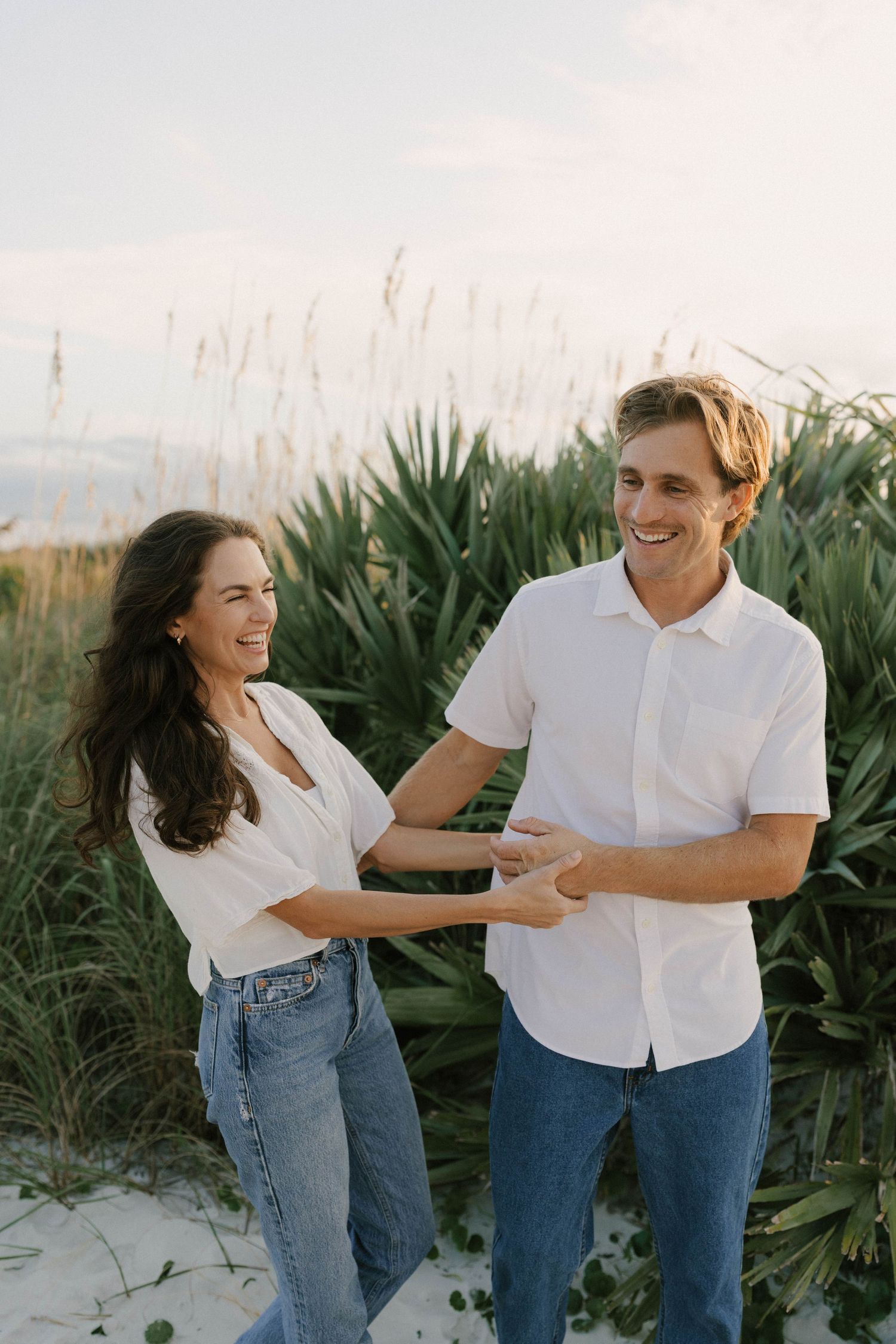 A smiling couple in white shirts and jeans hold hands on a sandy beach with sea oats and coastal plants behind them.
