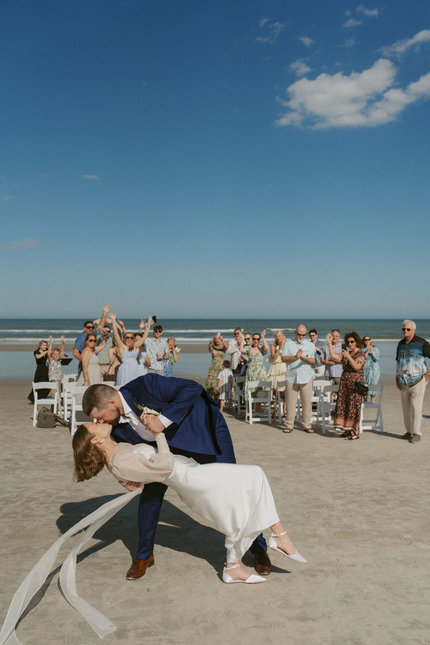 Couple performs romantic dip kiss on beach during wedding ceremony as guests cheer and take photos in background.