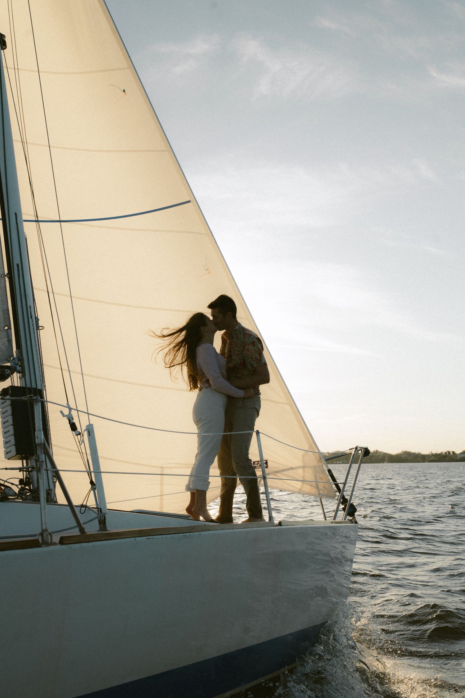 Silhouetted couple embraces on sailboat deck against sunset sky with white sail and calm waters in background.