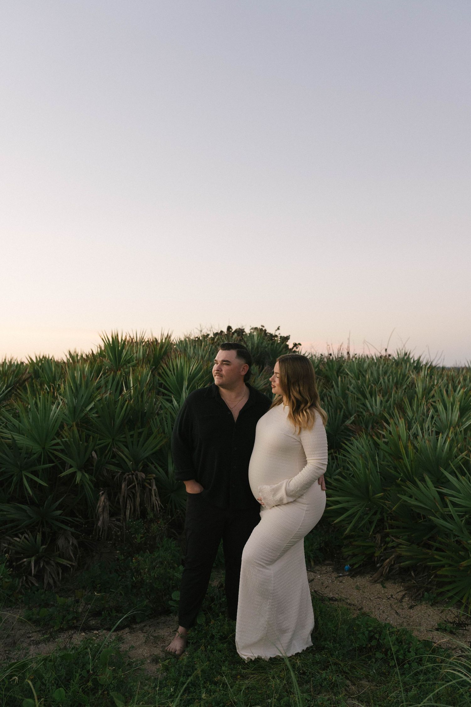 A couple embraces in a romantic moment at sunset among tall green plants in a field.