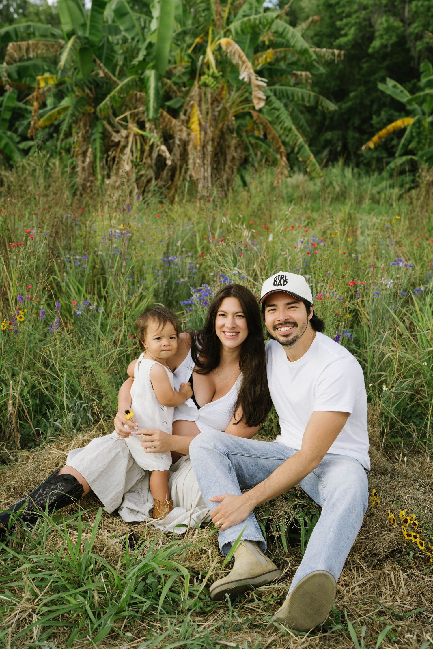 Family wearing casual white outfits sits together in a grassy field with wildflowers on a sunny afternoon.