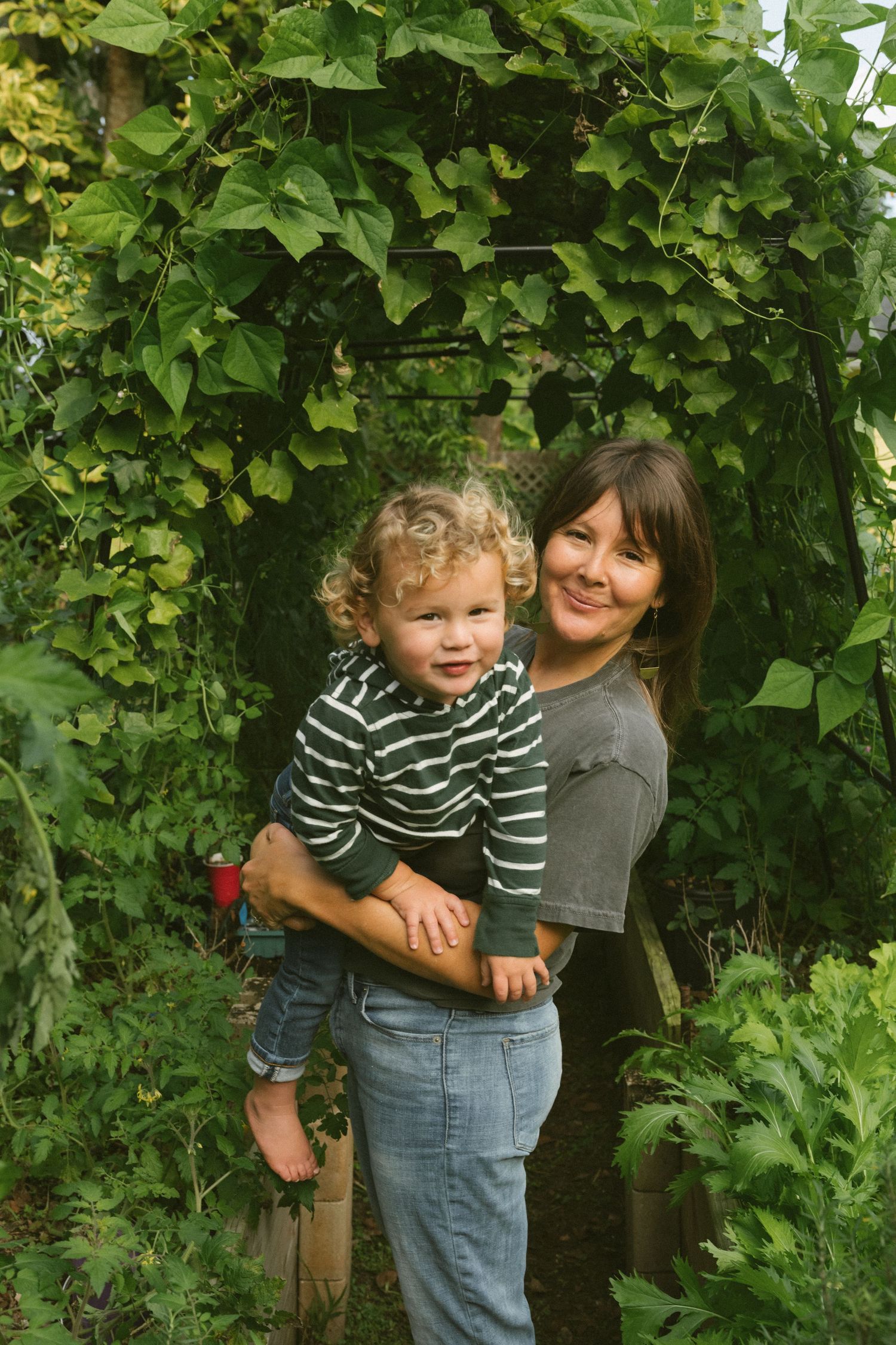 A small toddler in a striped sweater stands with an adult among lush green foliage and vines in a garden setting.