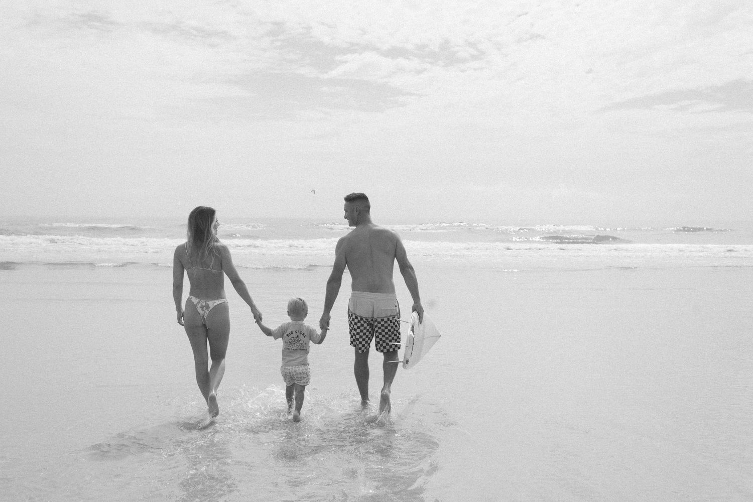 A family walks together holding hands along a misty beach shoreline in black and white.
