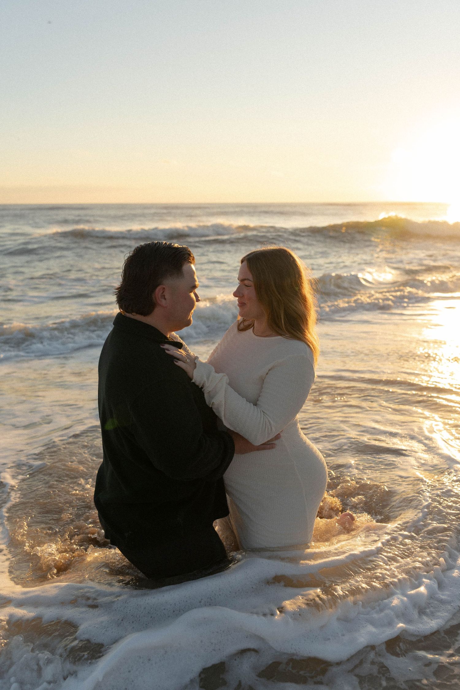 A couple sits together in the ocean surf at sunset while waves crash around them on the beach.
