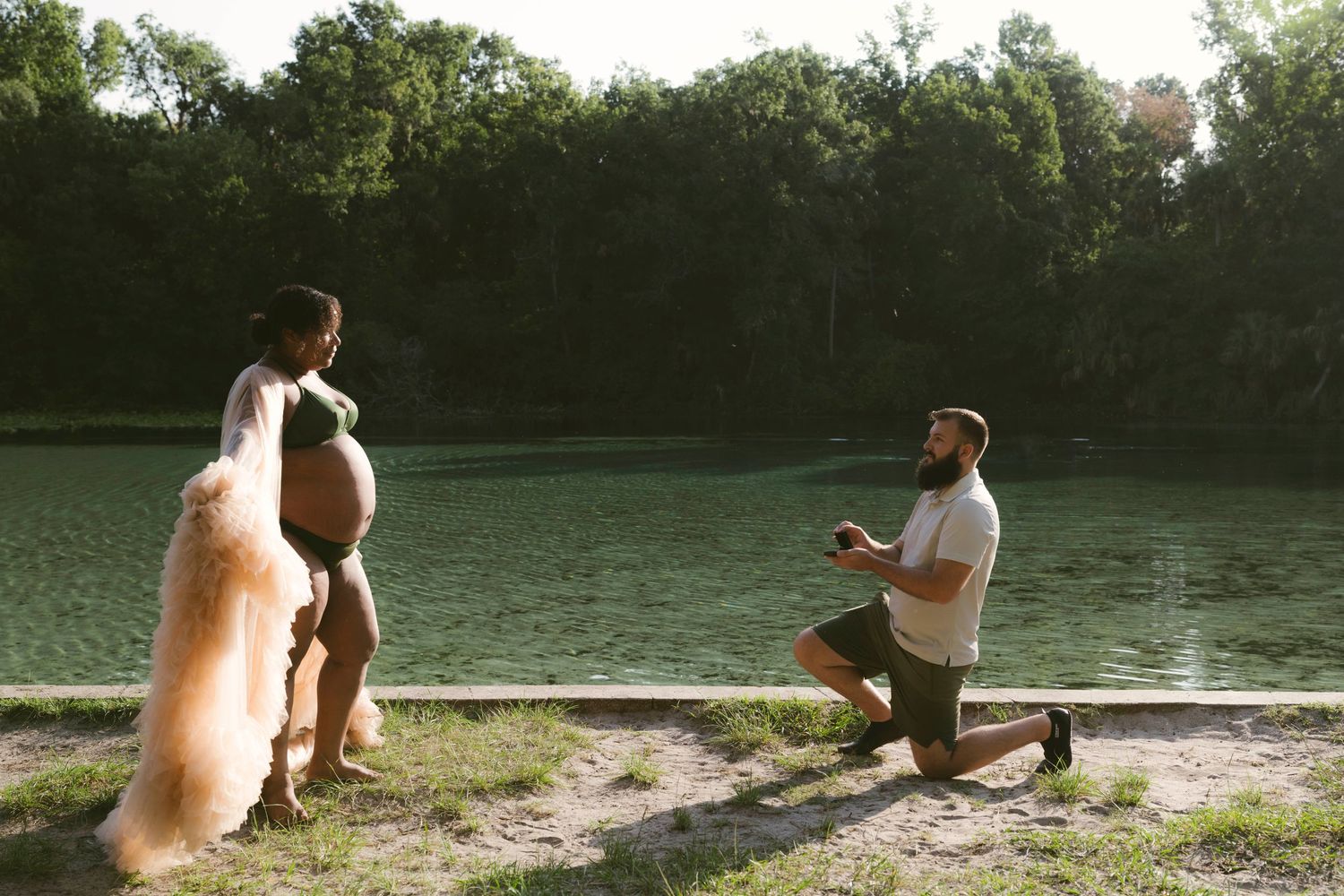 A proposal scene by a lake at sunset with two dogs watching as someone kneels down on one knee.