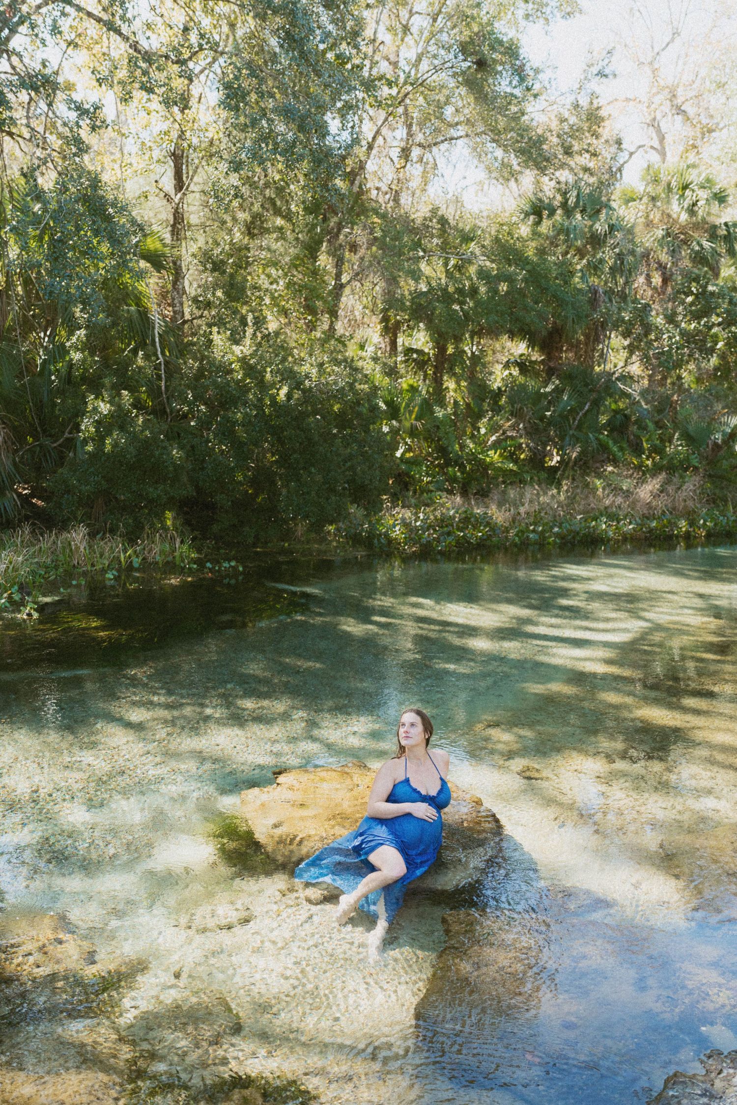 Someone relaxes on a rock in a serene natural spring surrounded by lush trees and crystal clear waters.