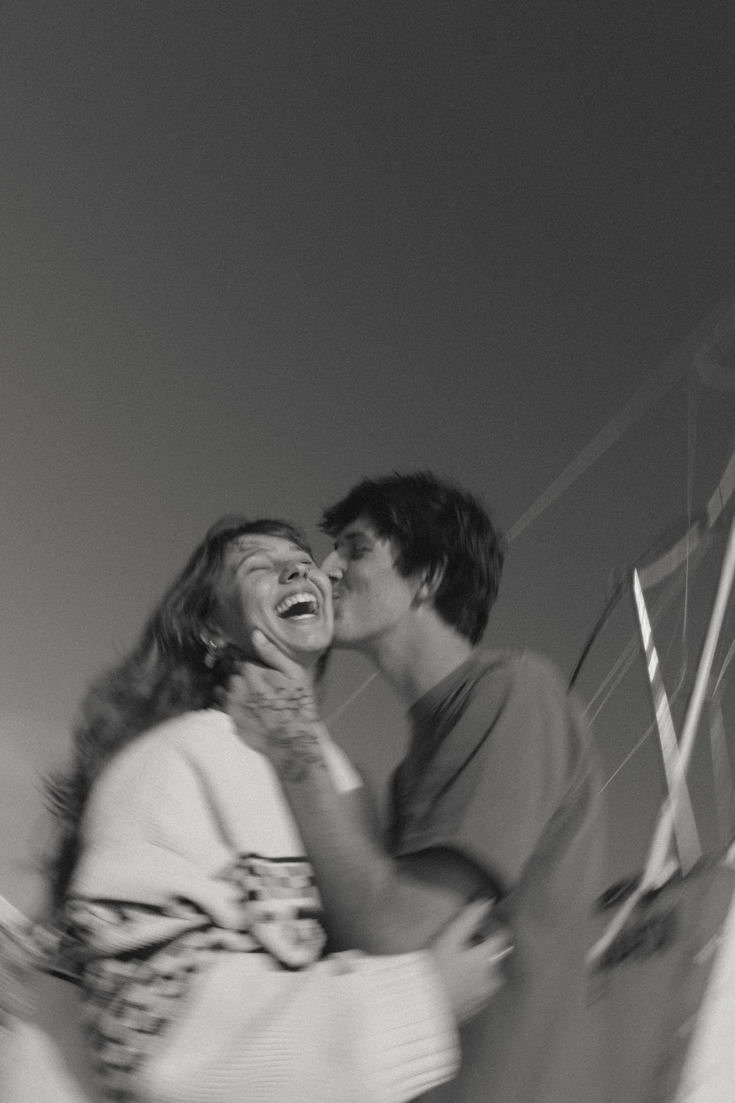 Black and white photo of a joyful couple, woman laughing as man kisses her cheek outdoors.