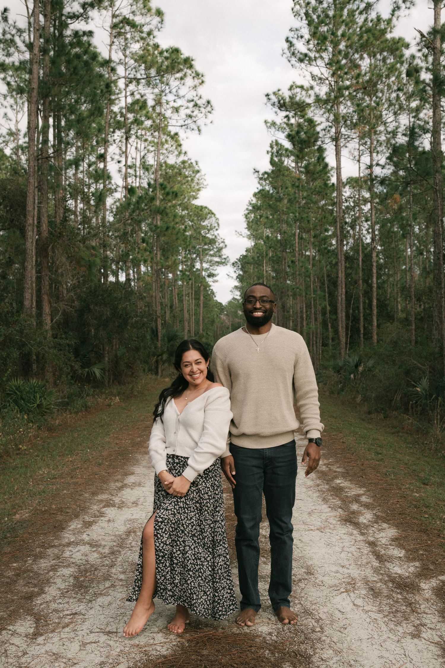 A couple stands together on a dirt path surrounded by tall pine trees on an overcast day.