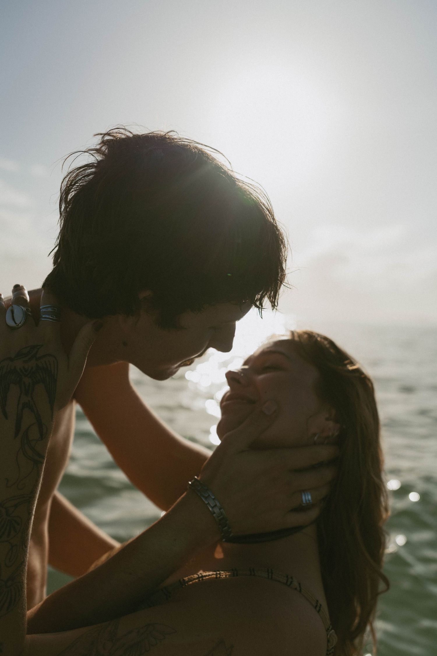 A couple shares a romantic moment by the ocean, with sunlight glowing behind them as he gently holds her face.