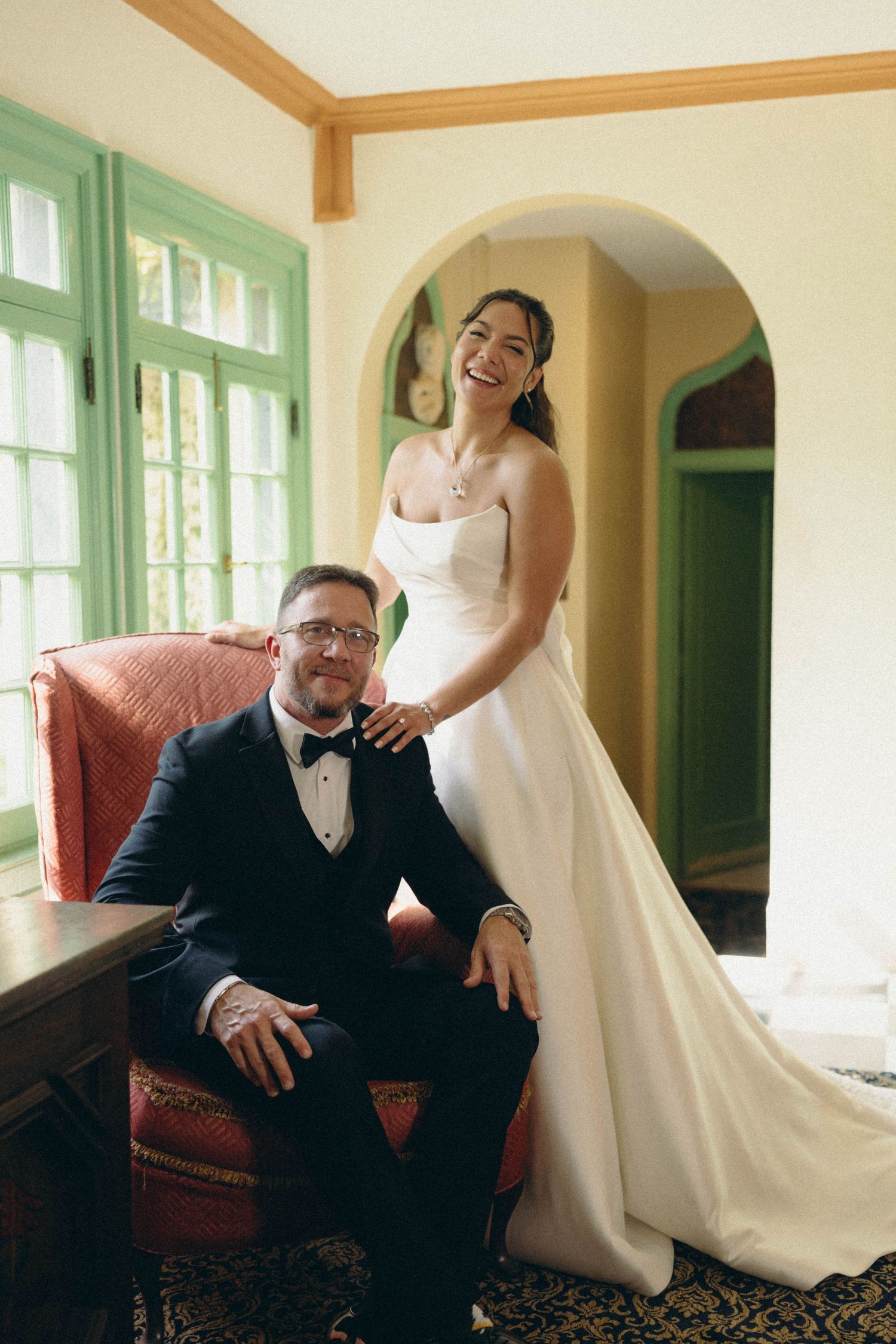 Wedding portrait of a couple by window featuring groom seated in leather chair and bride in white gown in sunlit room.