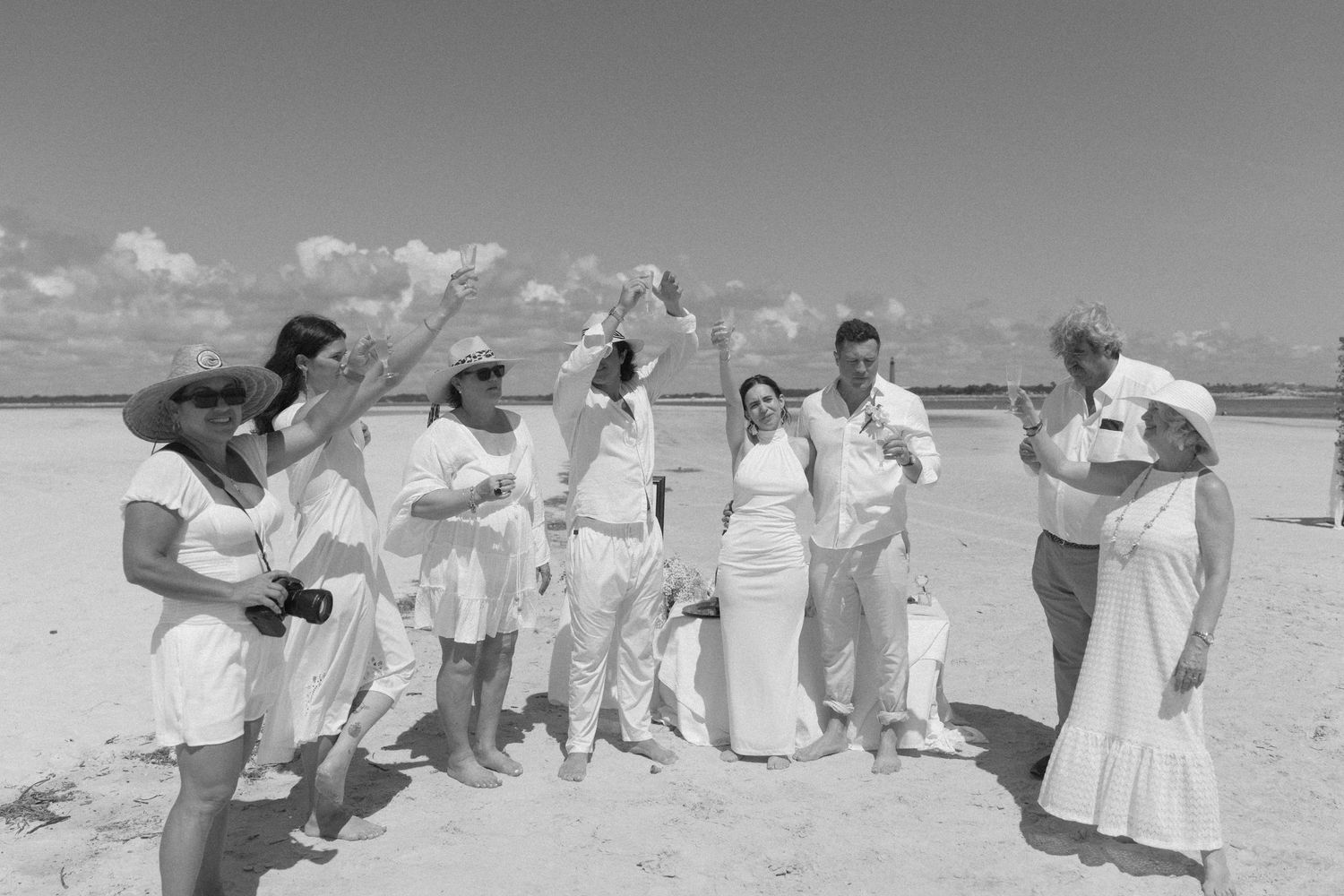 Group of people in white clothing dancing and celebrating on a beach in a black and white photograph.