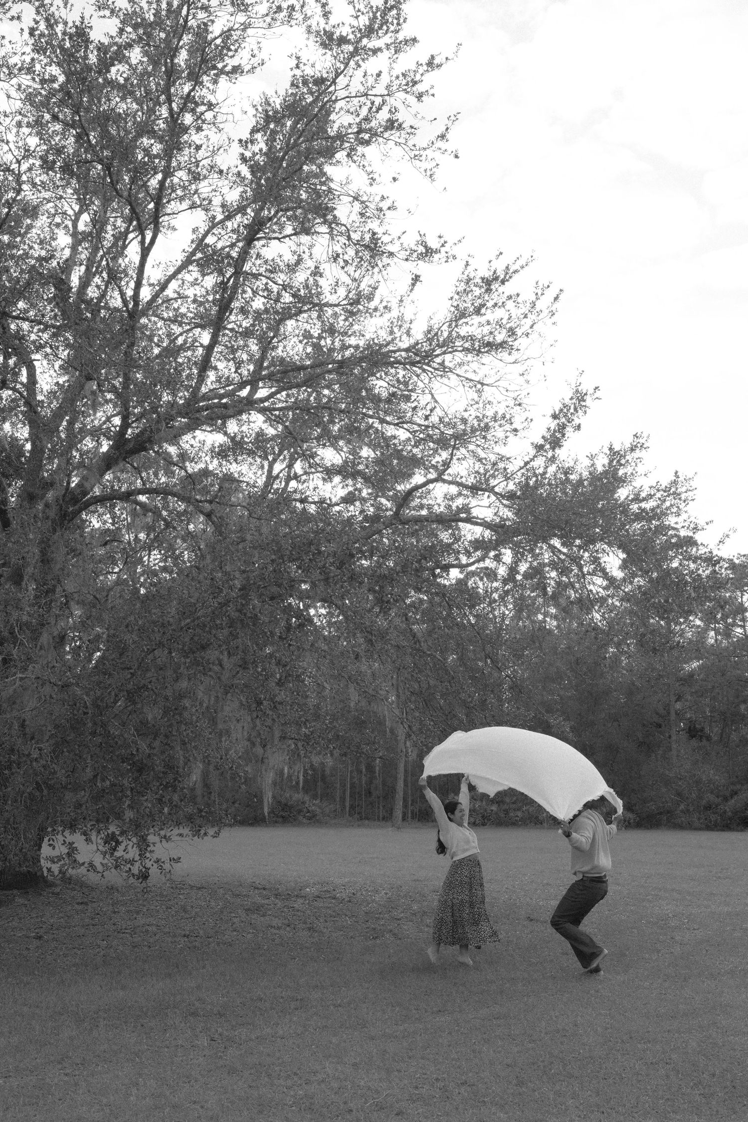 Two figures playing with a white sheet together in a field with trees in the background, captured in black and white.