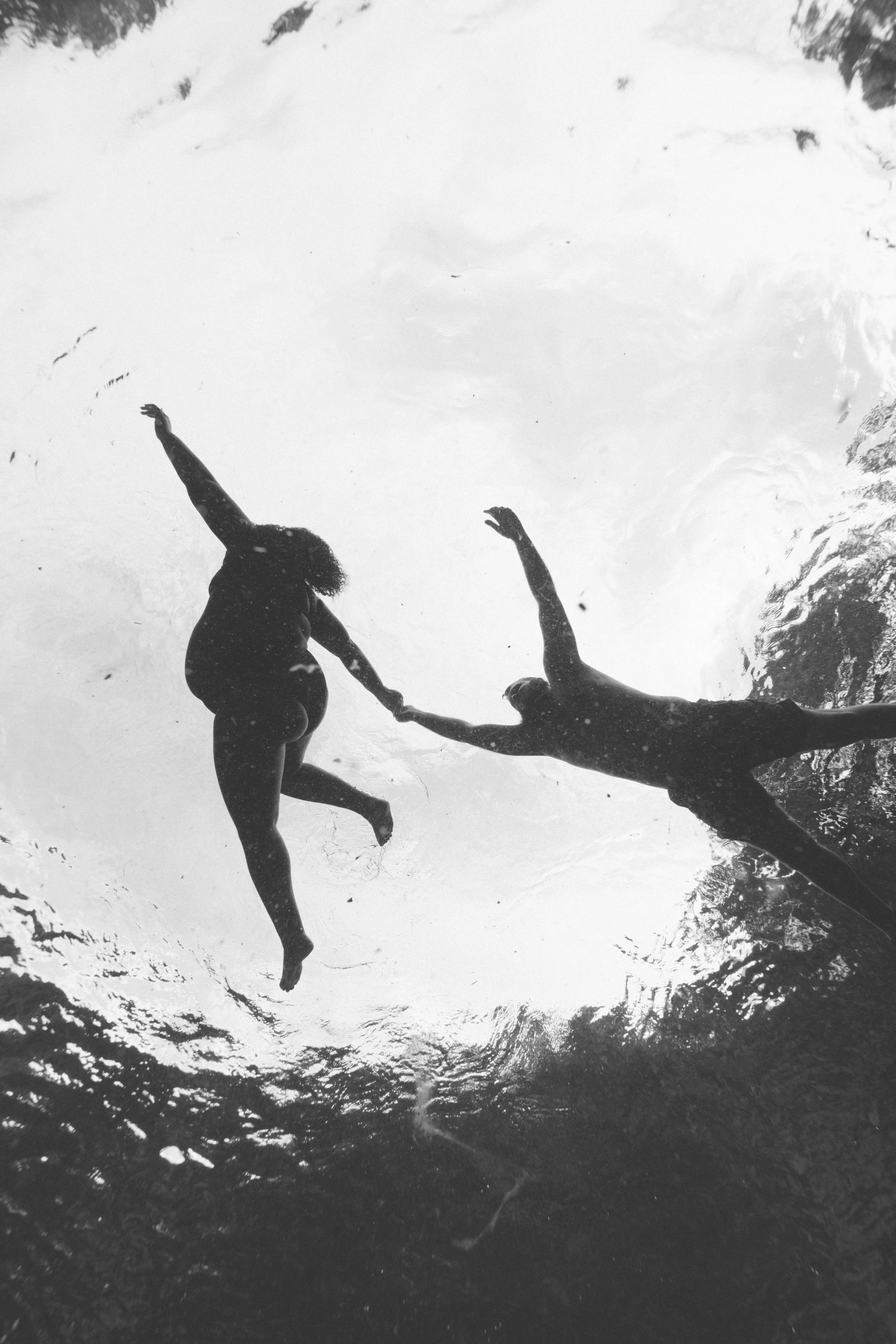 A black and white underwater view of silhouetted divers leaping into dark water in sequence against a bright sky backdrop.