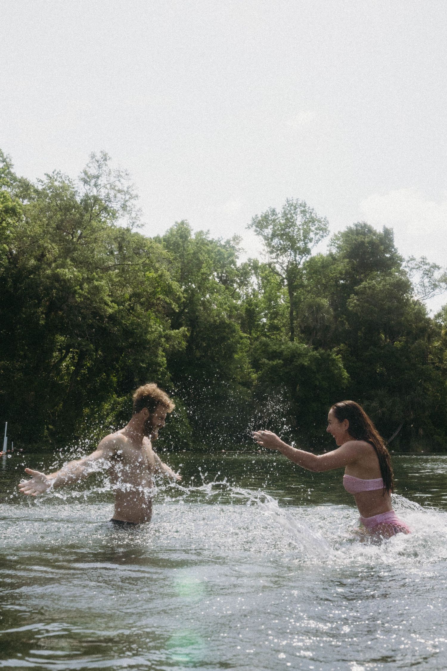Two people playing and splashing in a river on a summer day with trees lining the banks in the background.