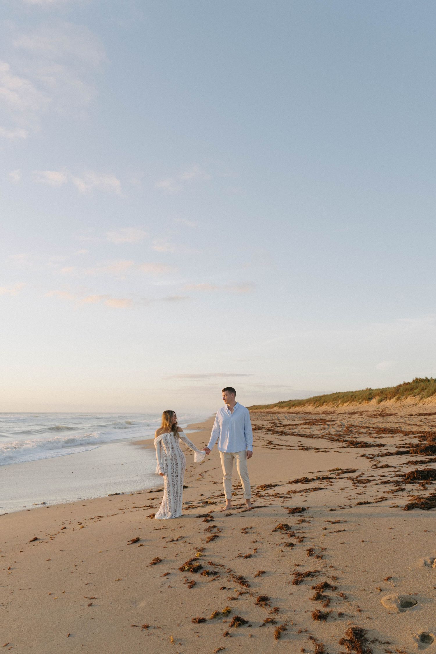 Couple holding hands walking on a sandy beach at sunset, wearing white outfits with ocean waves in the background.