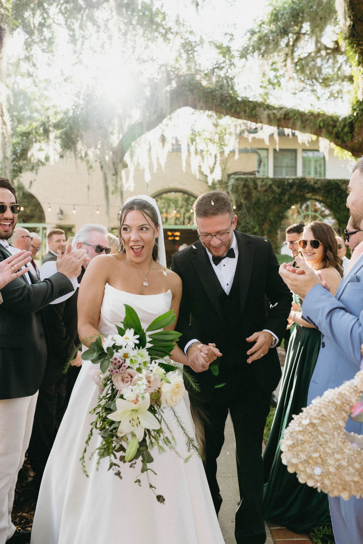 At an outdoor wedding ceremony under a large oak tree, a couple celebrates with guests as they walk down the aisle.