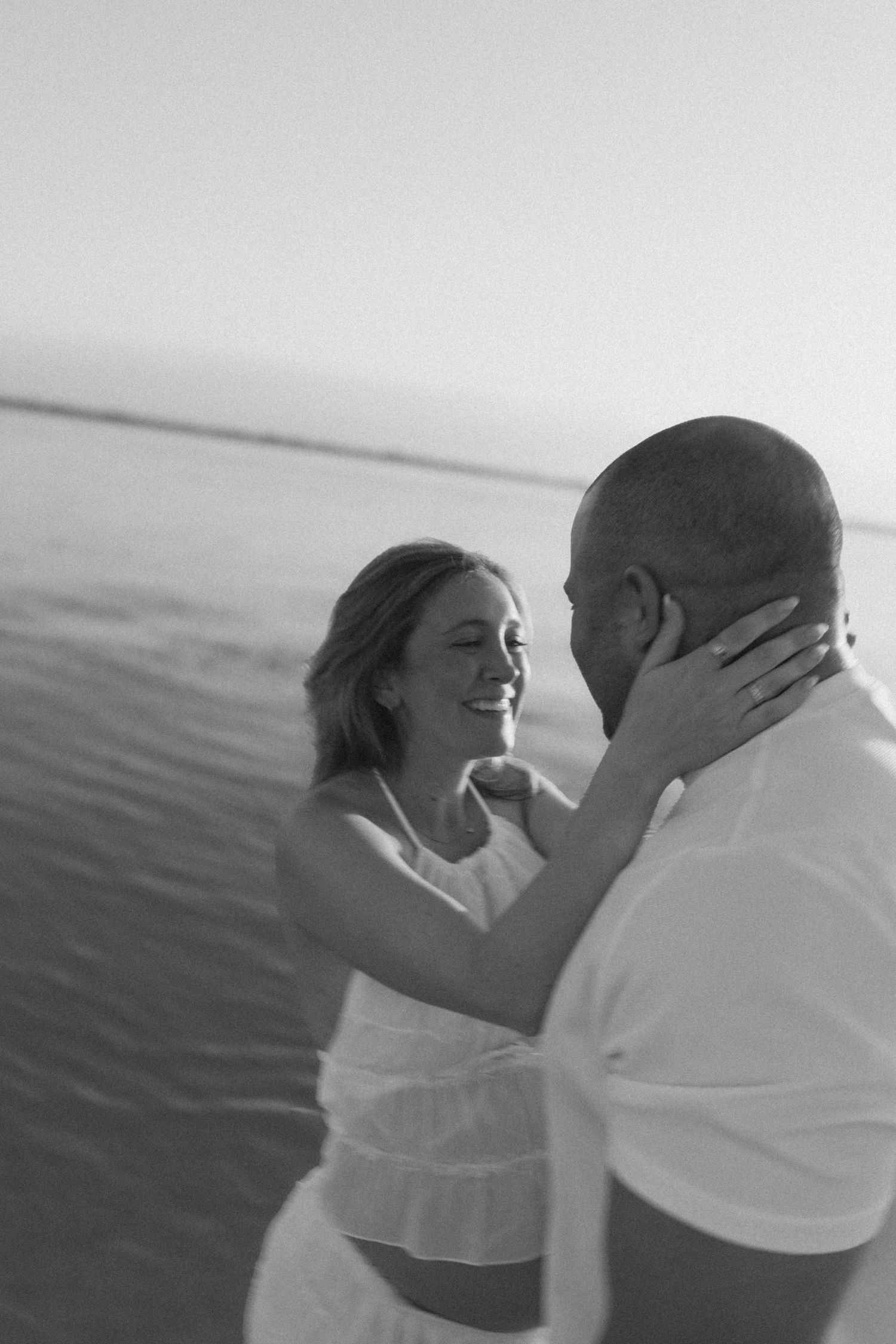 Black and white photo of a smiling couple embracing near the water, woman in white dress touching mans face.