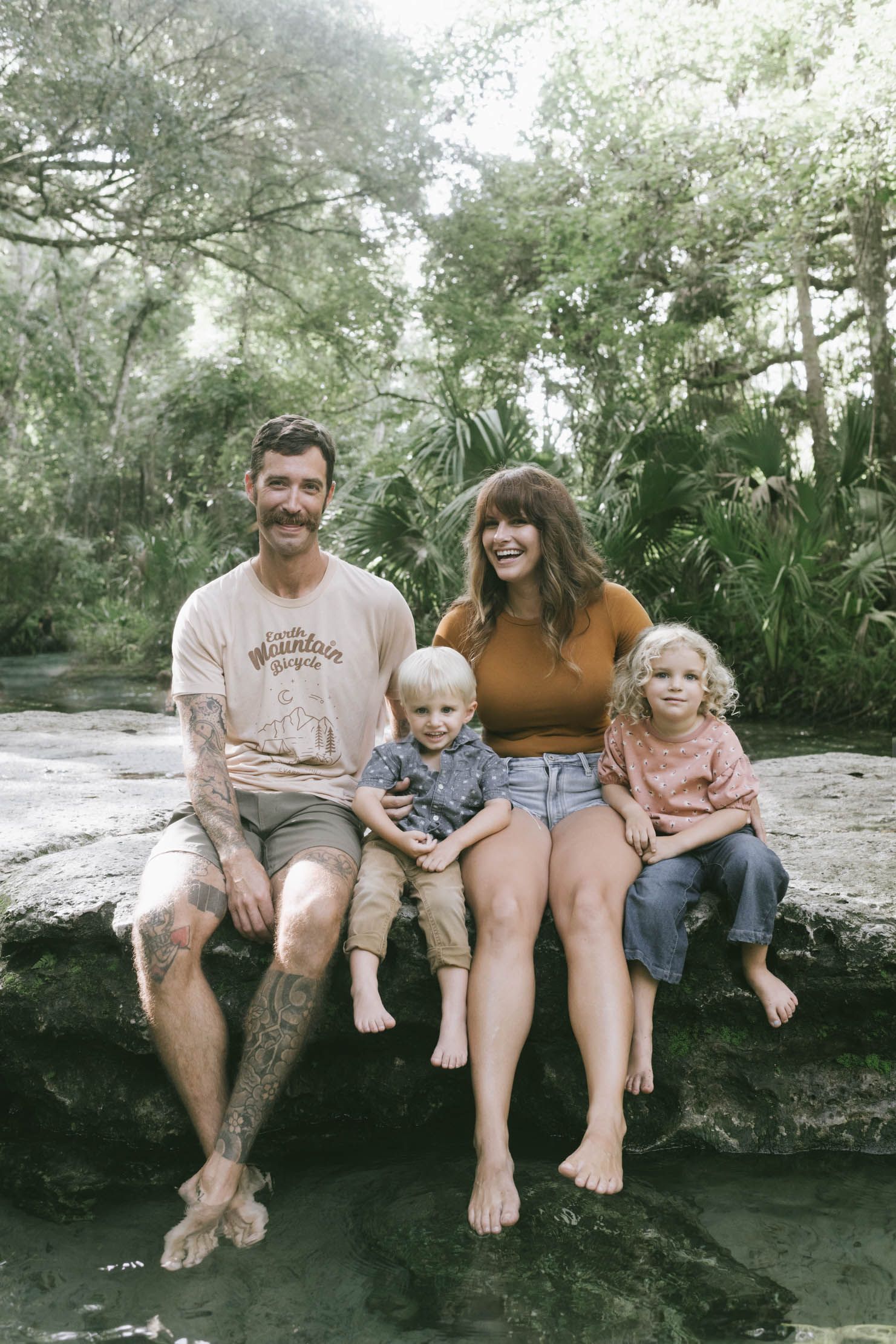 A family sits together barefoot on a dark rock formation surrounded by lush green foliage in a natural outdoor setting.