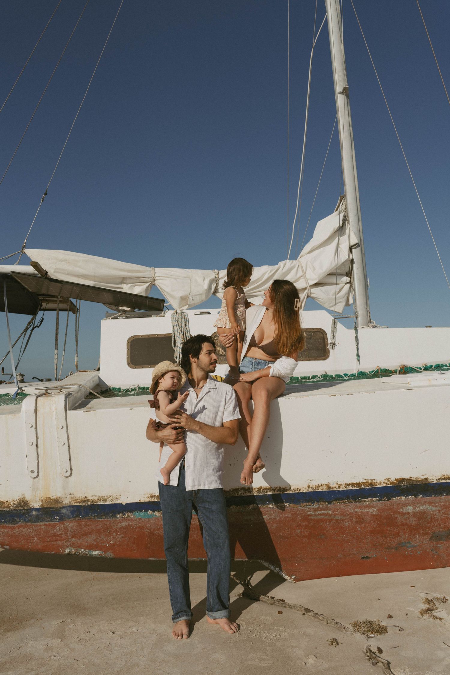 Family of four posing barefoot in front of a weathered white sailboat on a sunny beach day.
