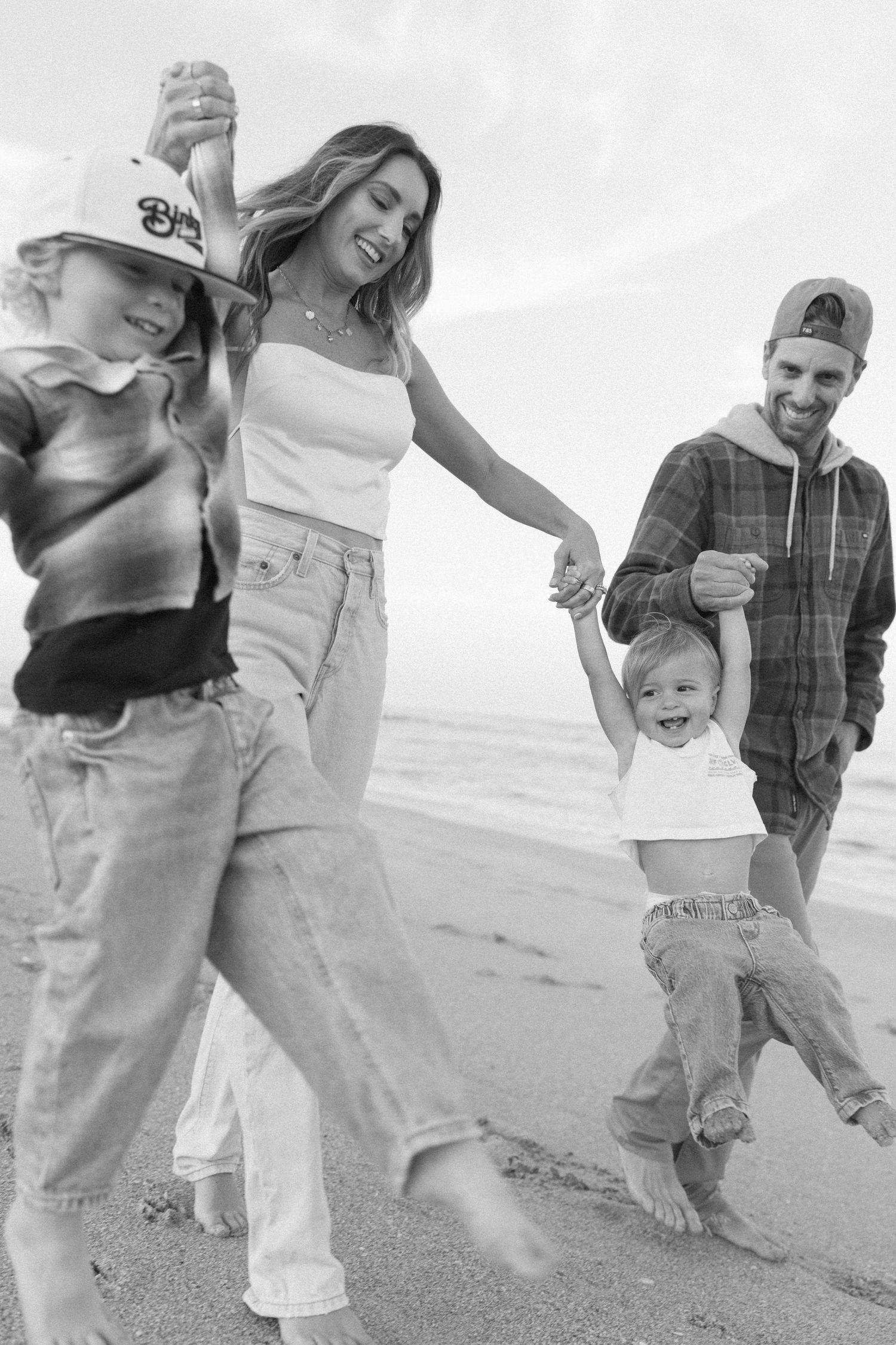 A family walks together playfully on a beach in black and white, showing their feet and legs in casual attire.