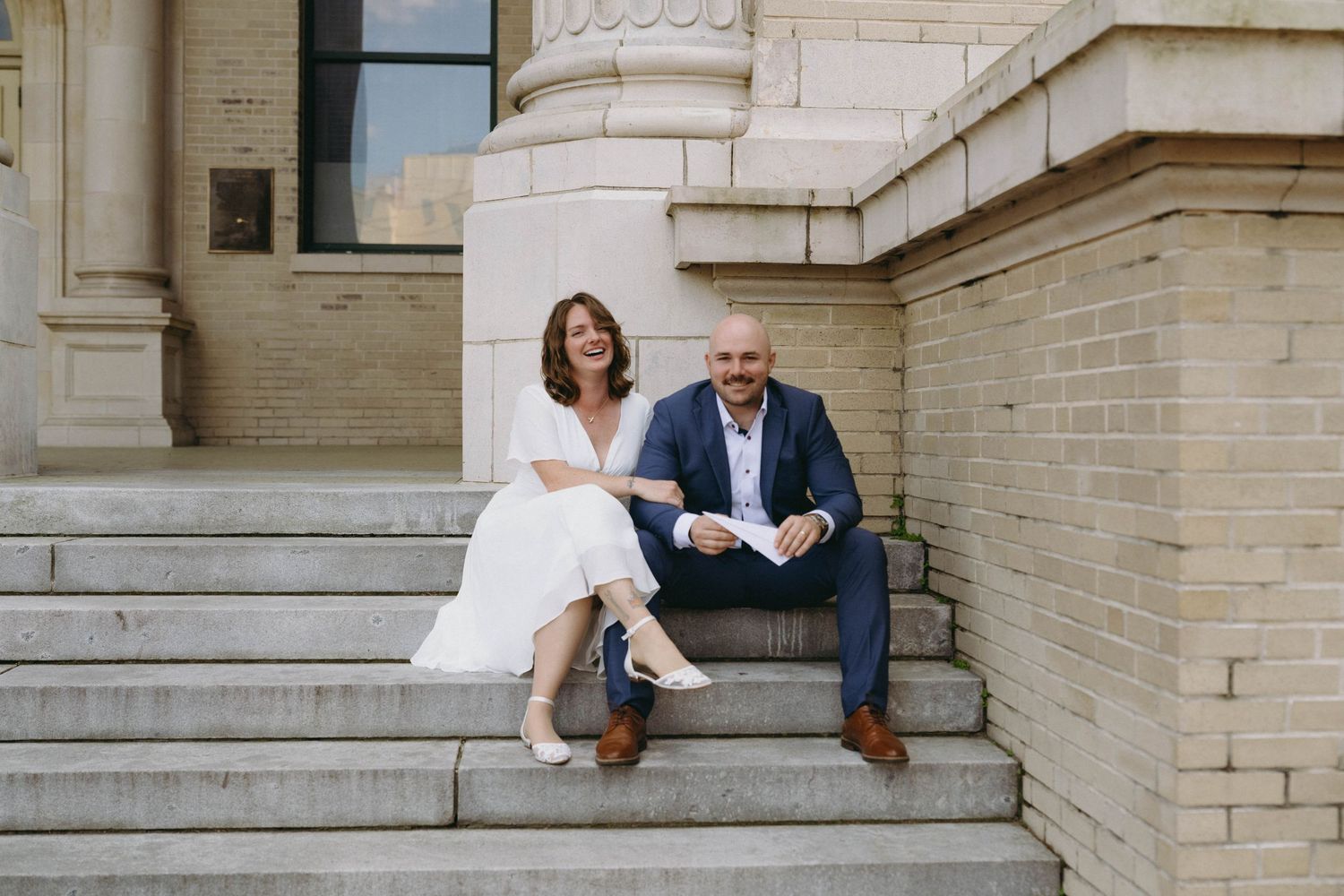A couple sits together on outdoor stone steps in casual attire during a sunny day.