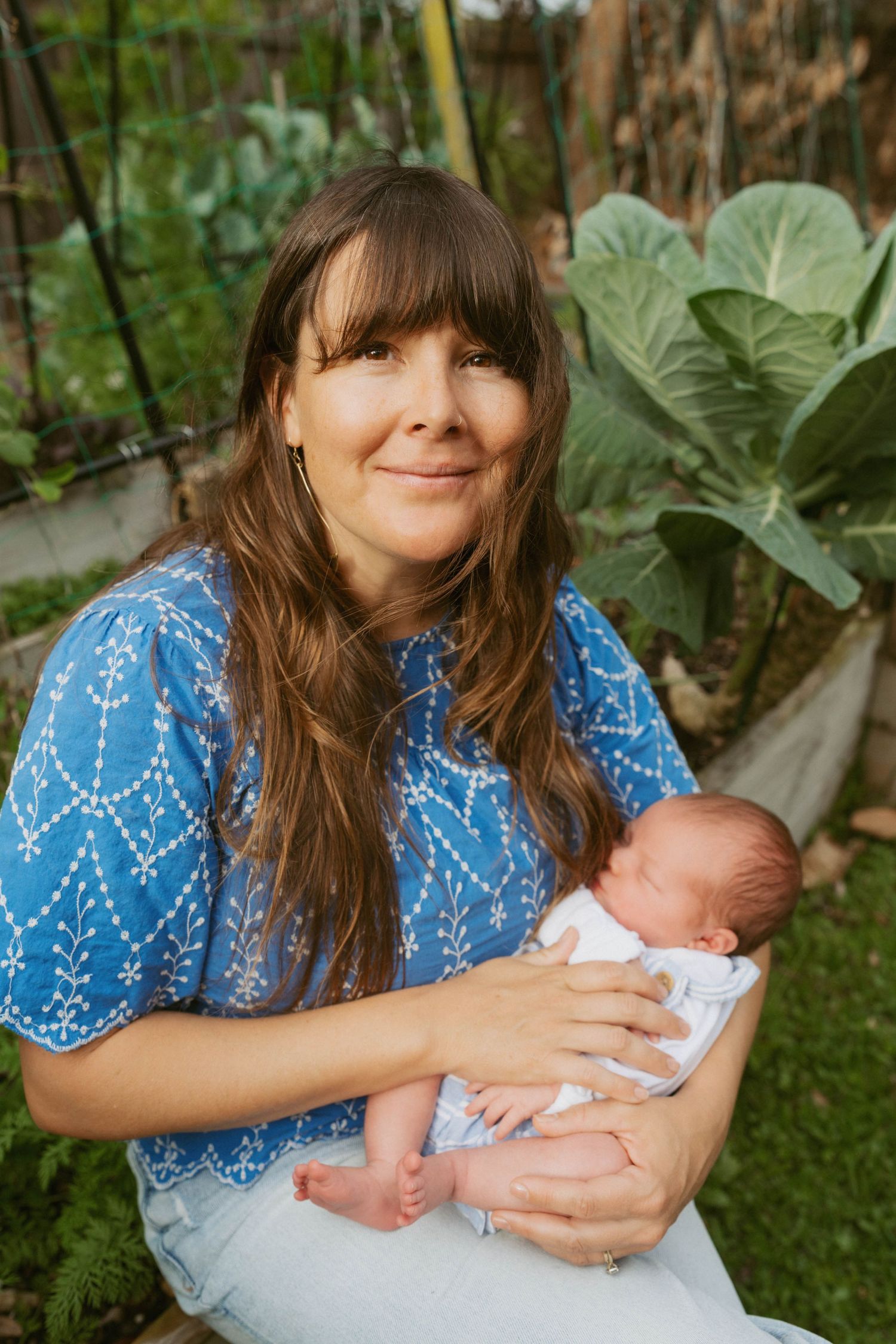 Woman in blue dress holding newborn baby in outdoor garden with green cabbage plants in background.