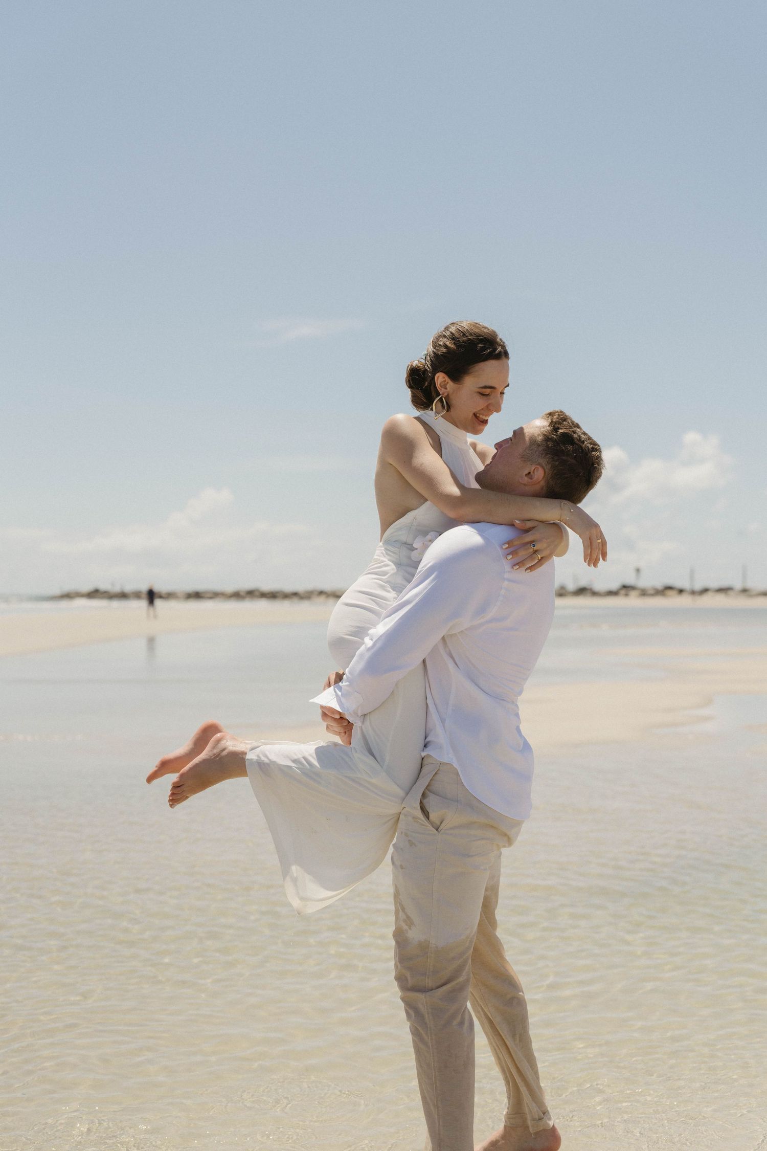 A couple in white embraces on a sunny beach as they share a romantic moment by the ocean.