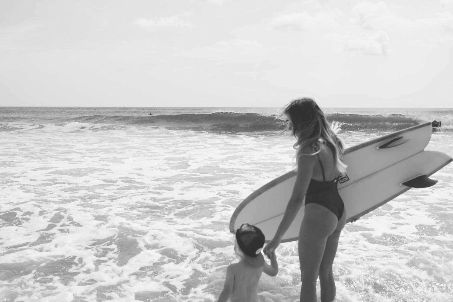 A surfer in a swimsuit stands at the ocean's edge with their surfboard while waves crash on the beach.